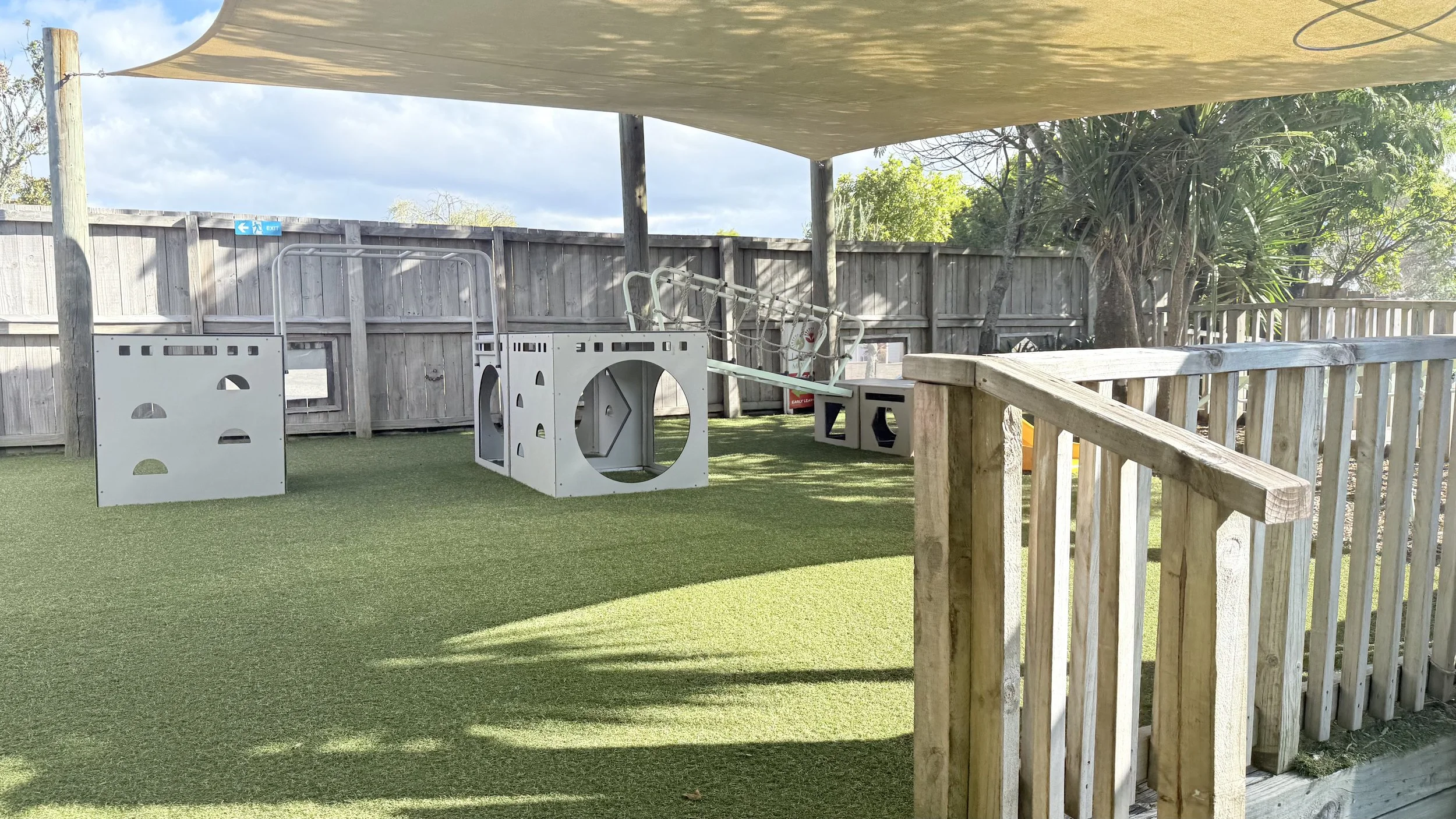 Empty outdoor children's play area with artificial grass, climbing structures, and a wooden fence, shaded by a canopy.