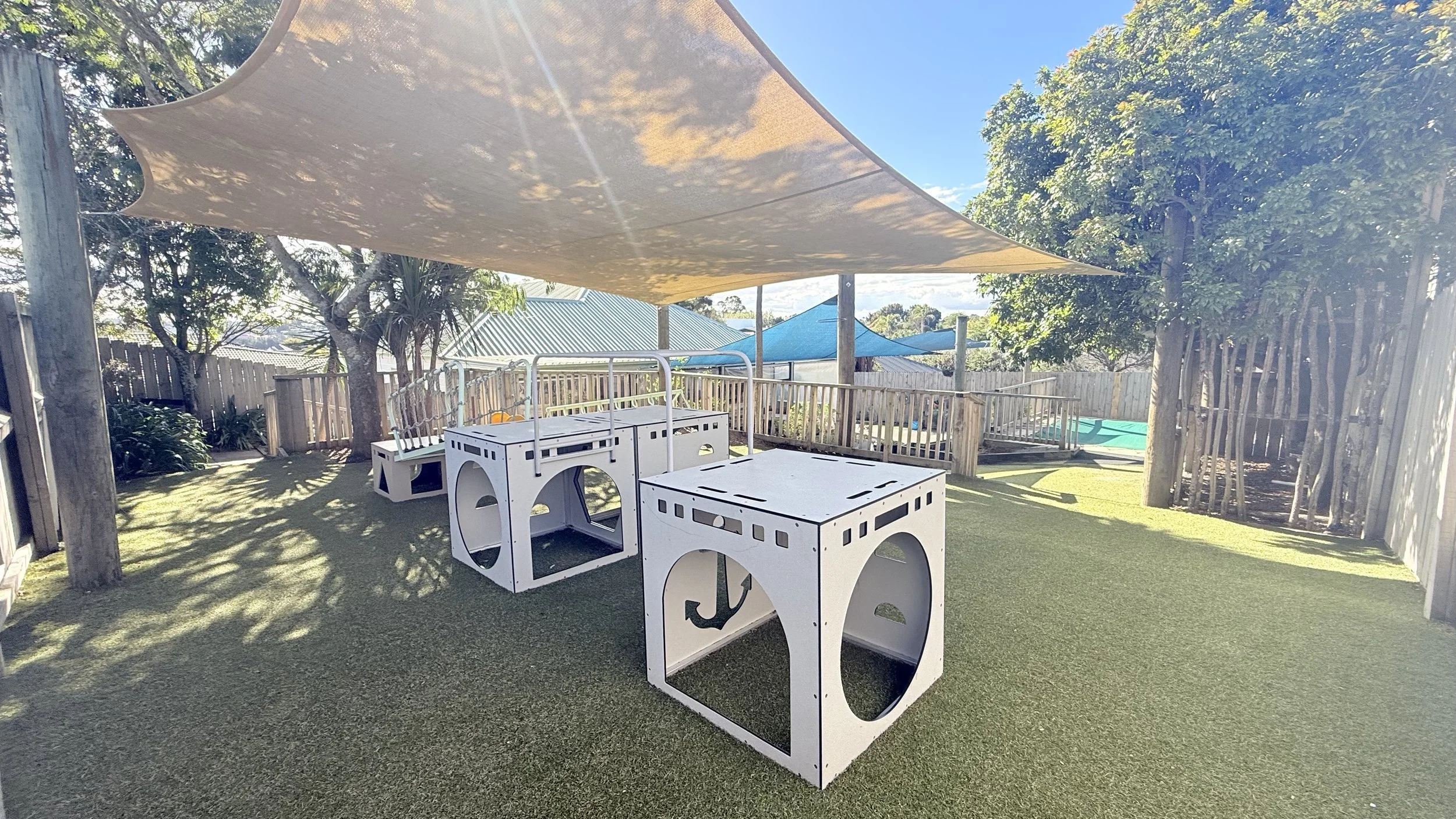 Outdoor playground with artificial grass, wooden fence, two large sail shades, and play structures shaped like nautical blocks with cutouts and black markings, under a sunny sky at Ponderosa Kids, Albany
