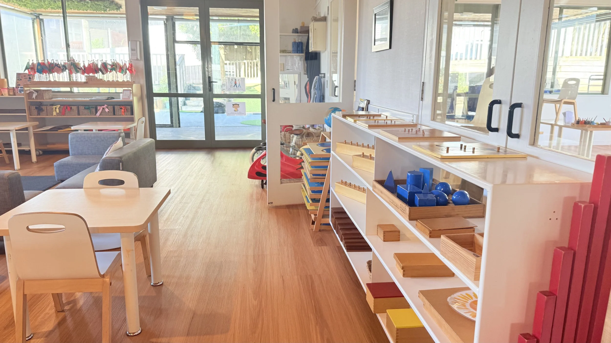 Interior of a classroom or playroom with wooden floors, white furniture, and shelves displaying educational toys and materials. Large windows and glass doors provide natural light and a view outside.