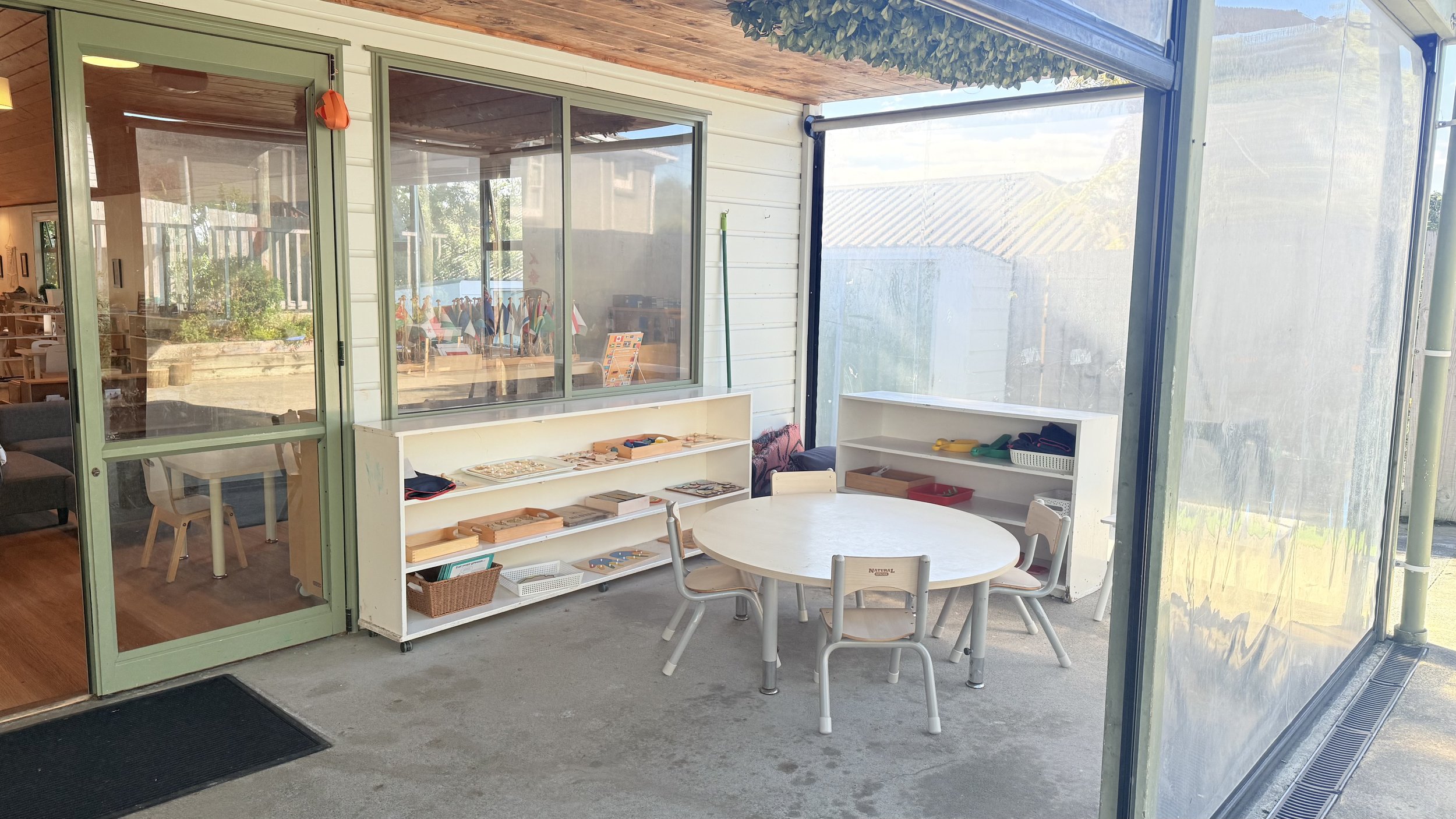 Outdoor classroom with a round table, chairs, and white storage shelves filled with educational materials, enclosed with transparent screens.