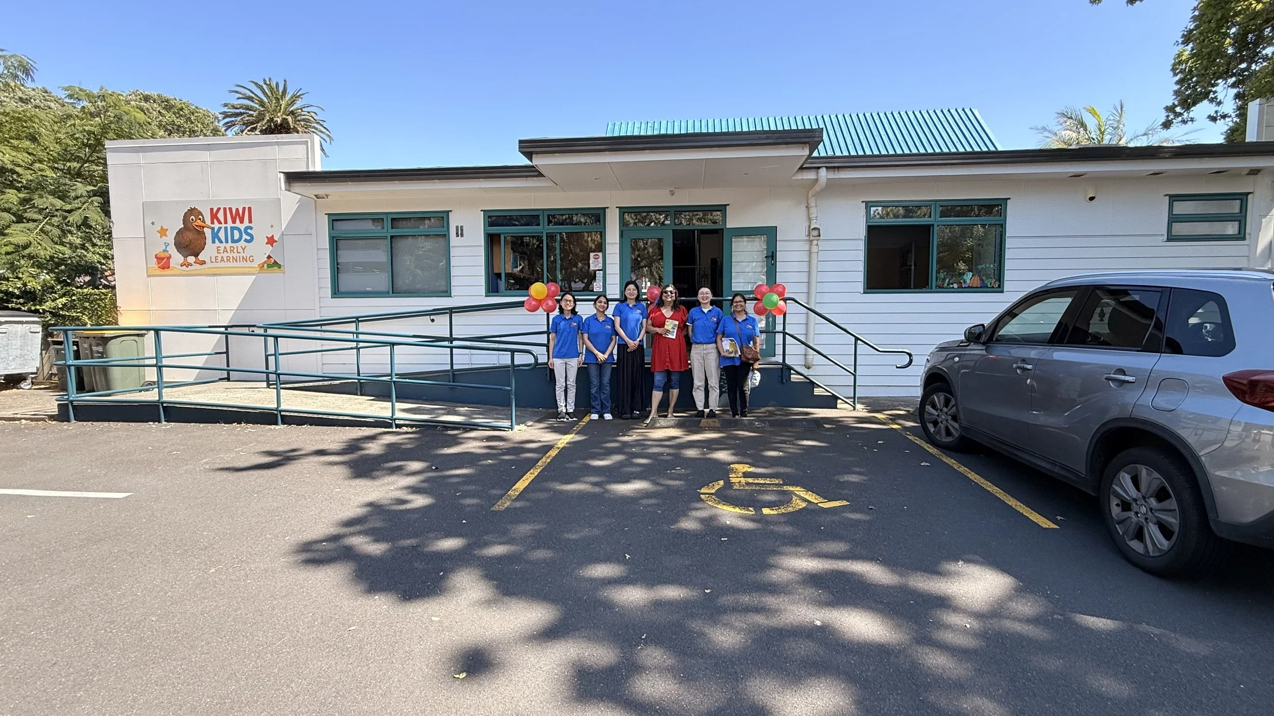 The Kiwi Kids team standing in front of Kiwi Kids Early Learning centre, with balloons and cheerful expressions, parked cars nearby, on a bright sunny day.