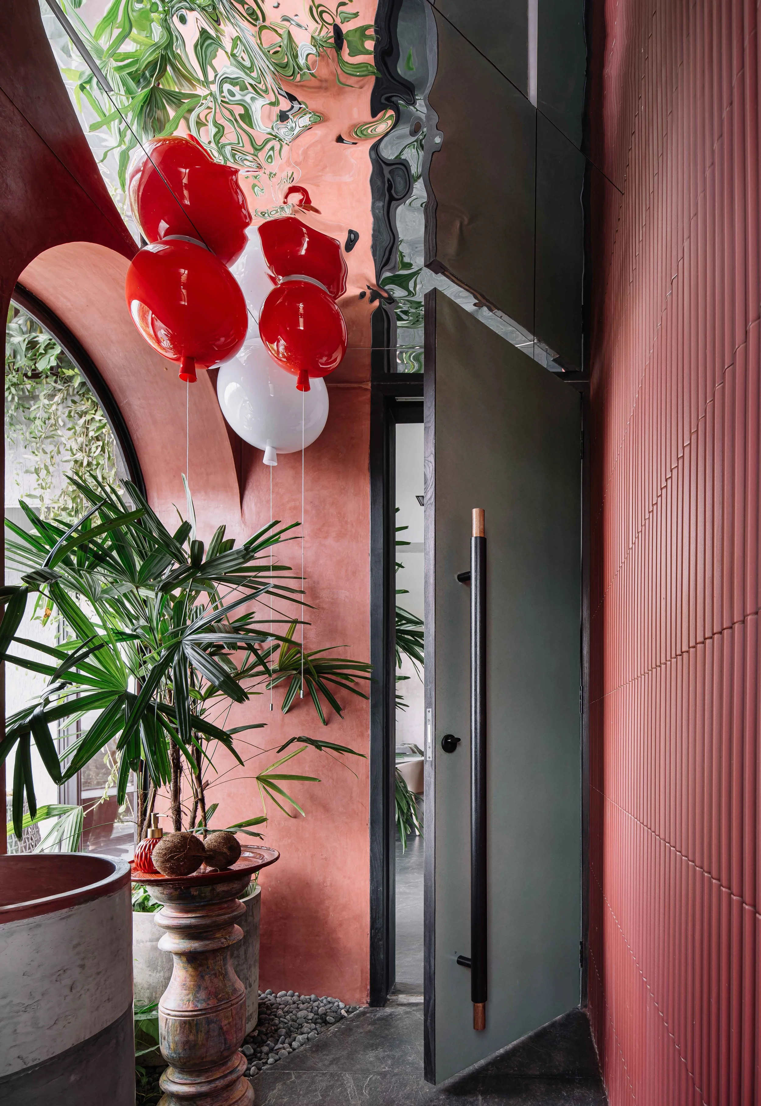 Arched interior with terracotta walls, sculptural red pendant lights, lush palms, and a dark pivot door at Gardens in the Sky, a Calicut residence designed by Workers of Art.