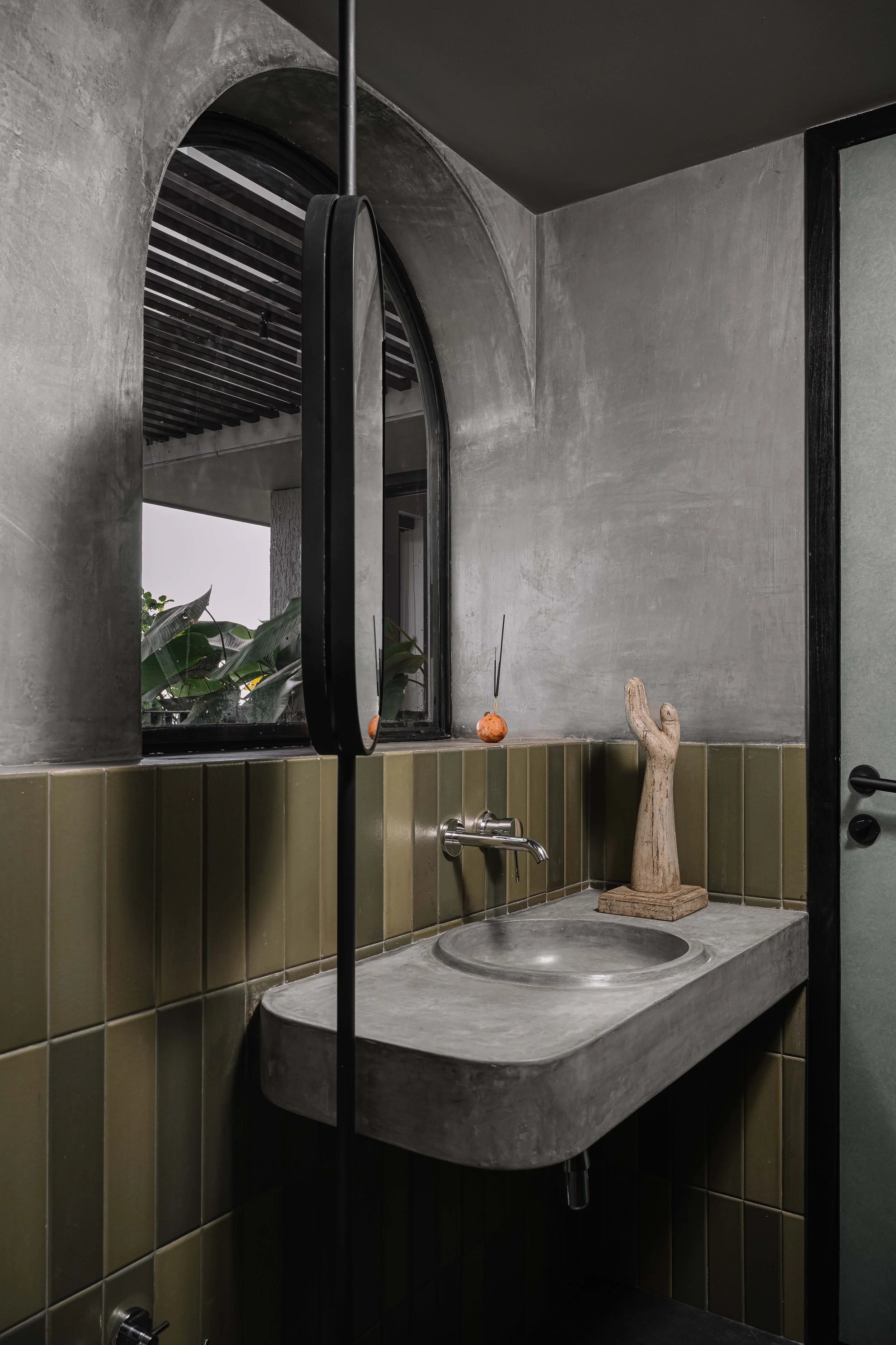 The guest bathroom features a cast-in-place washbasin and cement oxide finishes, complemented by an arched window that overlooks the landscaped balcony, bringing in natural light and reinforcing the sense of calm simplicity in the space.