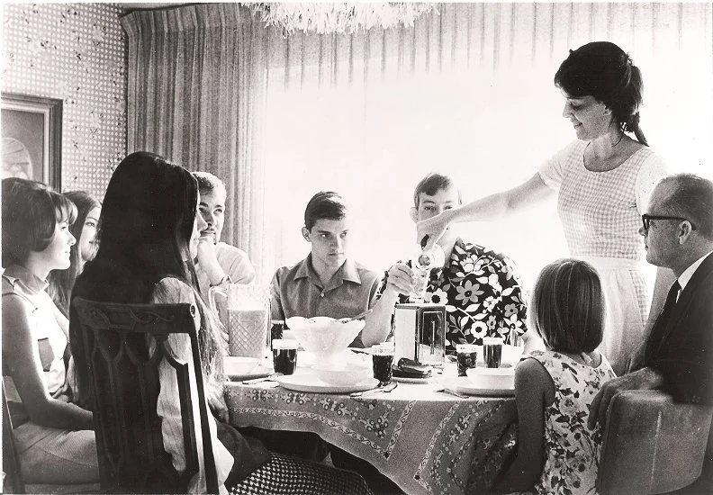 A woman pouring tea or coffee to a young girl at a family gathering around a table with children and adults in the background.