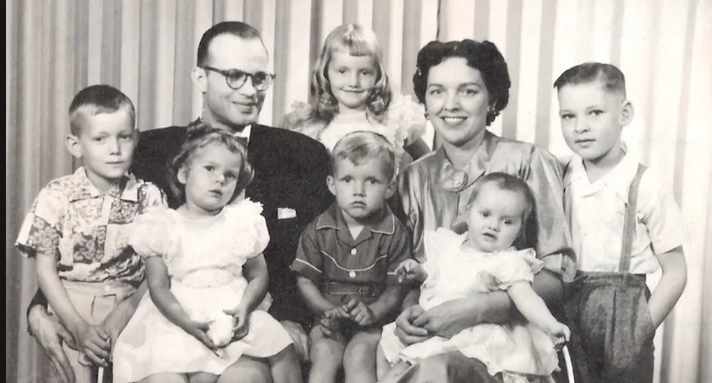 Black and white photo of a family group with two adults and six children, all dressed in vintage clothing, sitting and standing in front of a curtain background.