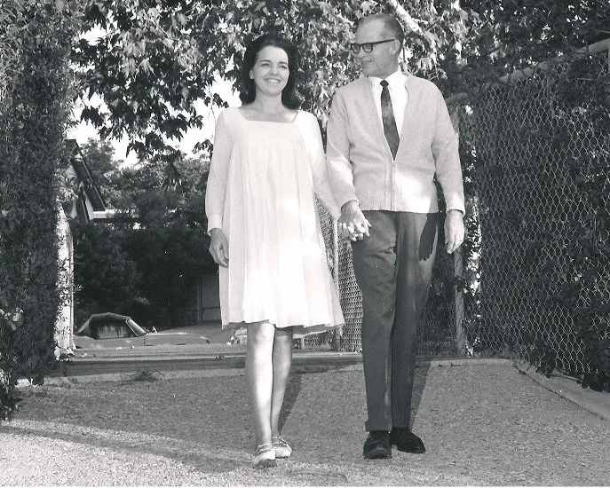 A young woman in a light-colored dress walking and holding hands with an older man in a suit outdoors on a sunny day.