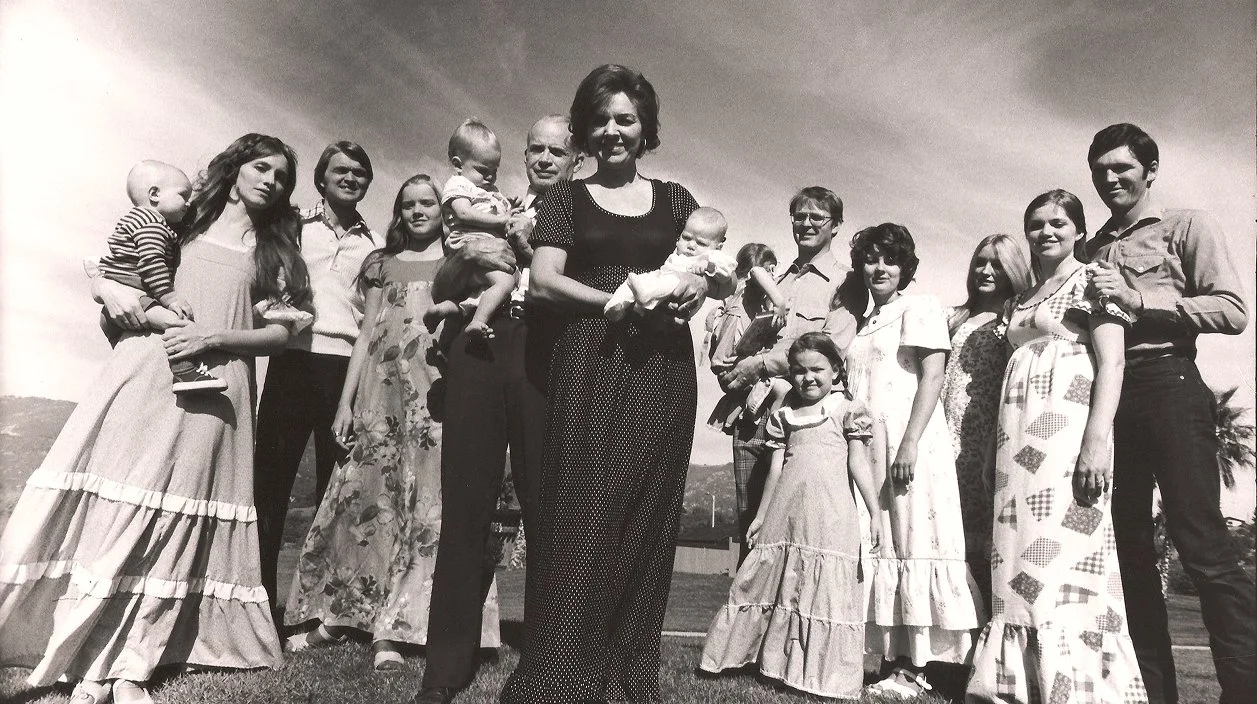 A black-and-white photo of a large family group outdoors standing on a grassy field, with several adults and children, some holding babies, posing for the camera.