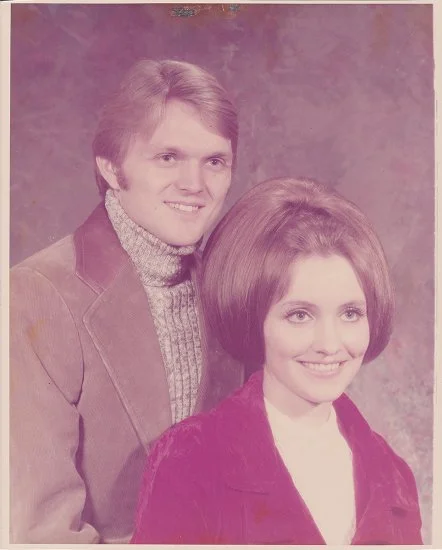 Vintage portrait of a young man and woman smiling, with the man standing behind the woman. The man wears a jacket and turtleneck, and the woman has a voluminous hairstyle and wears a jacket over a white top.