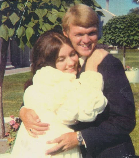A young man and woman hugging outdoors on a sunny day, smiling, with trees and a house in the background.