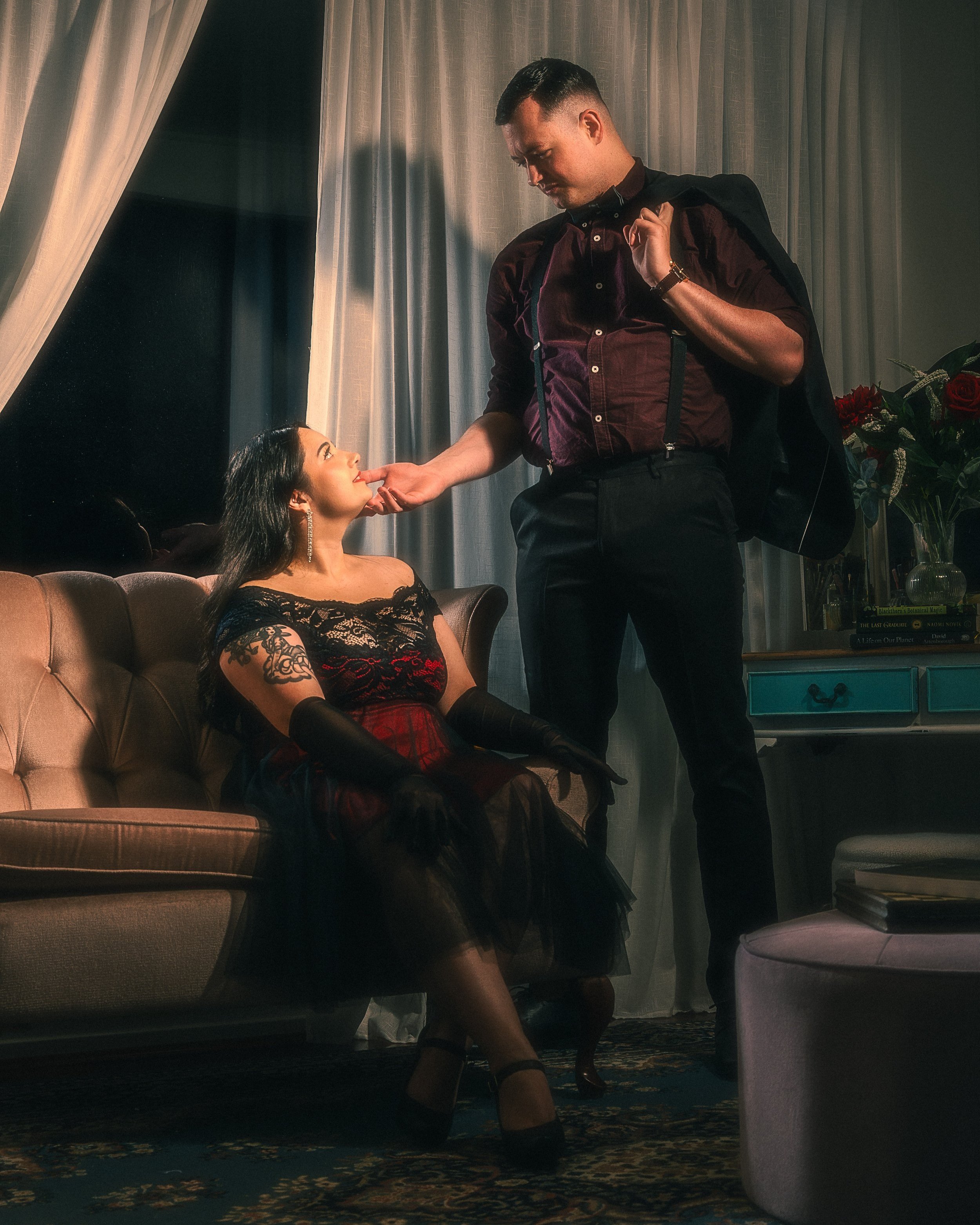 A man and woman in formal attire in a dimly lit room; the man stands and gently touches the woman's chin while she sits on a vintage sofa, looking up at him.