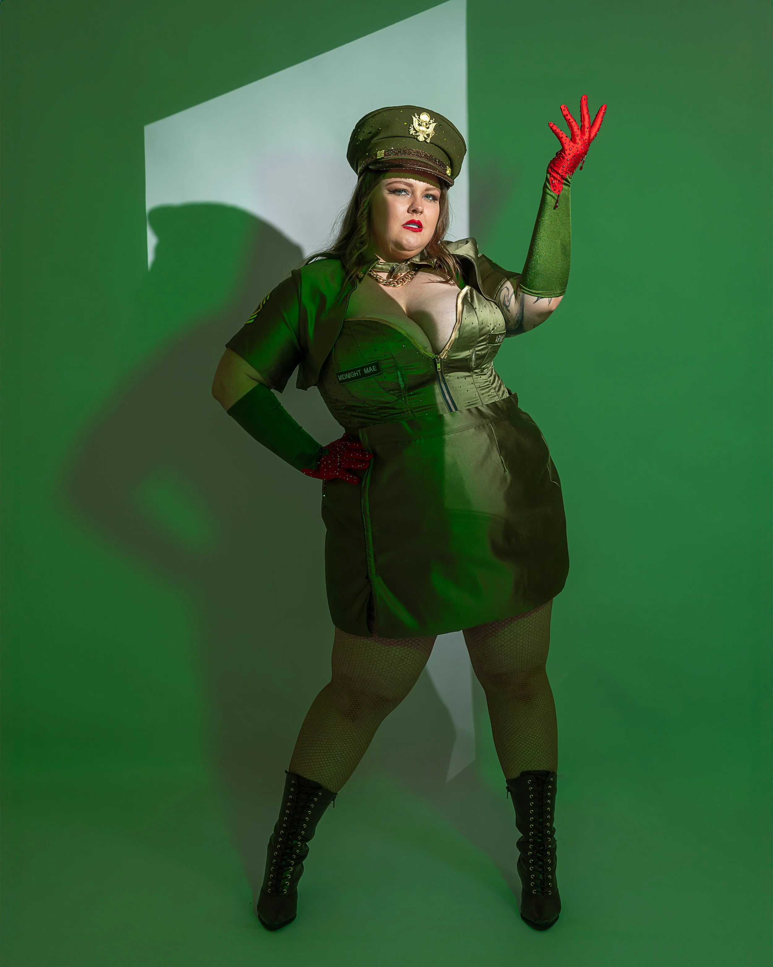 Woman dressed as a police officer with a military hat, posing with her right hand raised, wearing red gloves, fishnet stockings, and black high-heeled boots, standing against a green screen with a white projection.