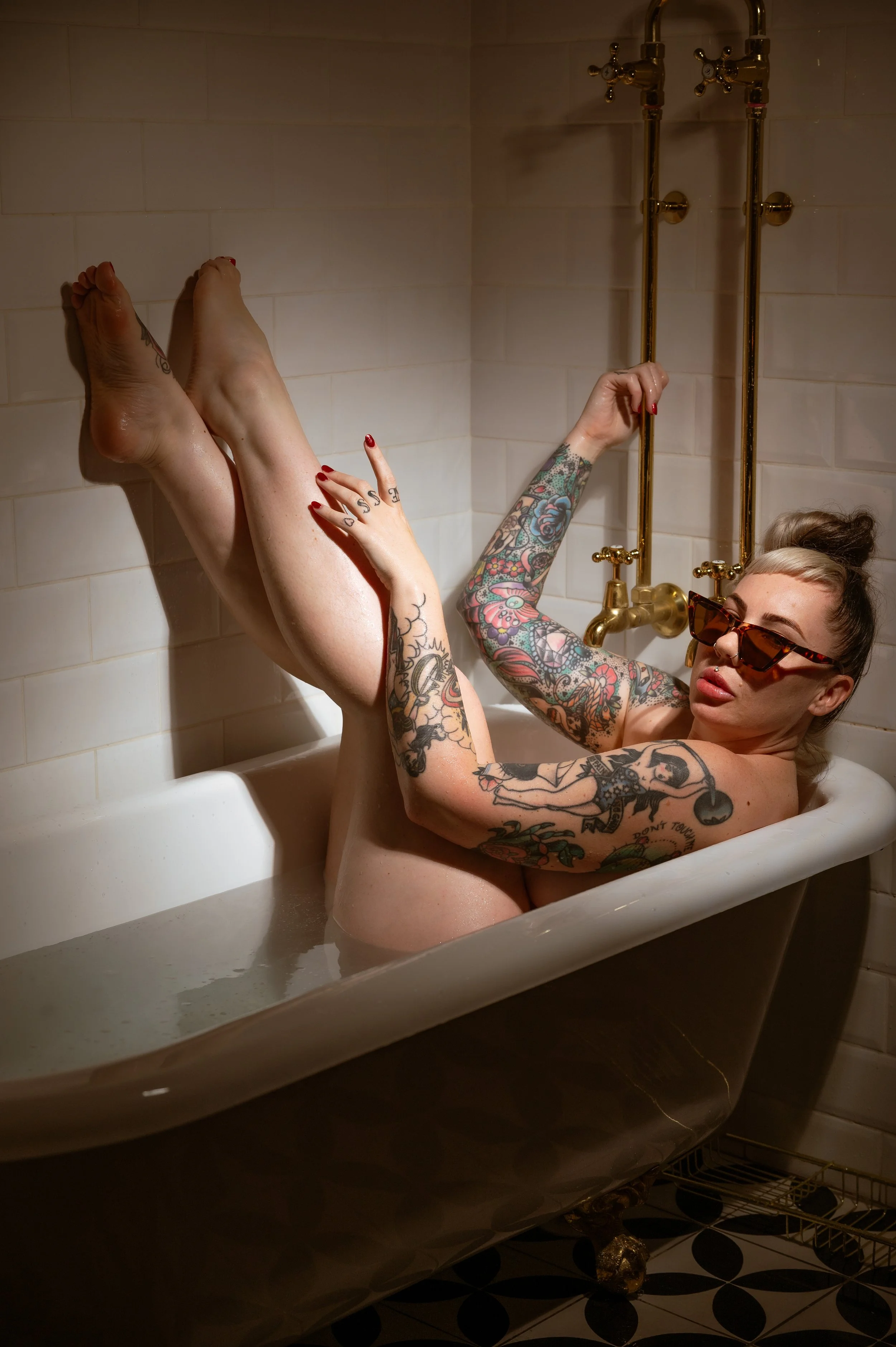 A tattooed woman wearing sunglasses relaxing in a vintage bathtub filled with water, her legs crossed and her right arm raised gripping the bath faucet.