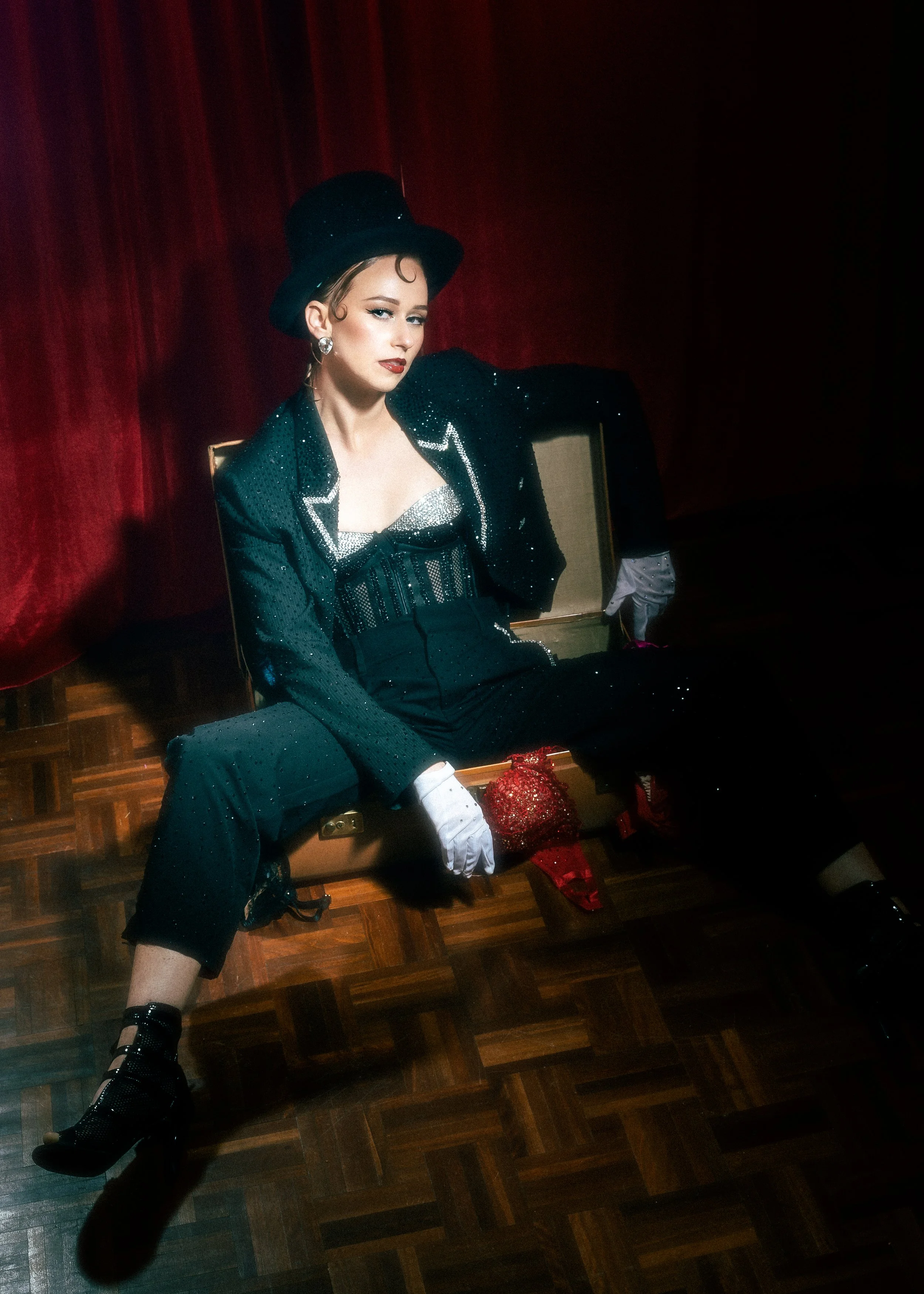A woman dressed in vintage black and silver clothing with a top hat, sitting on a beige suitcase in front of red velvet curtains, with accessories like gloves, earrings, and a clutch, in a theatrical setting.