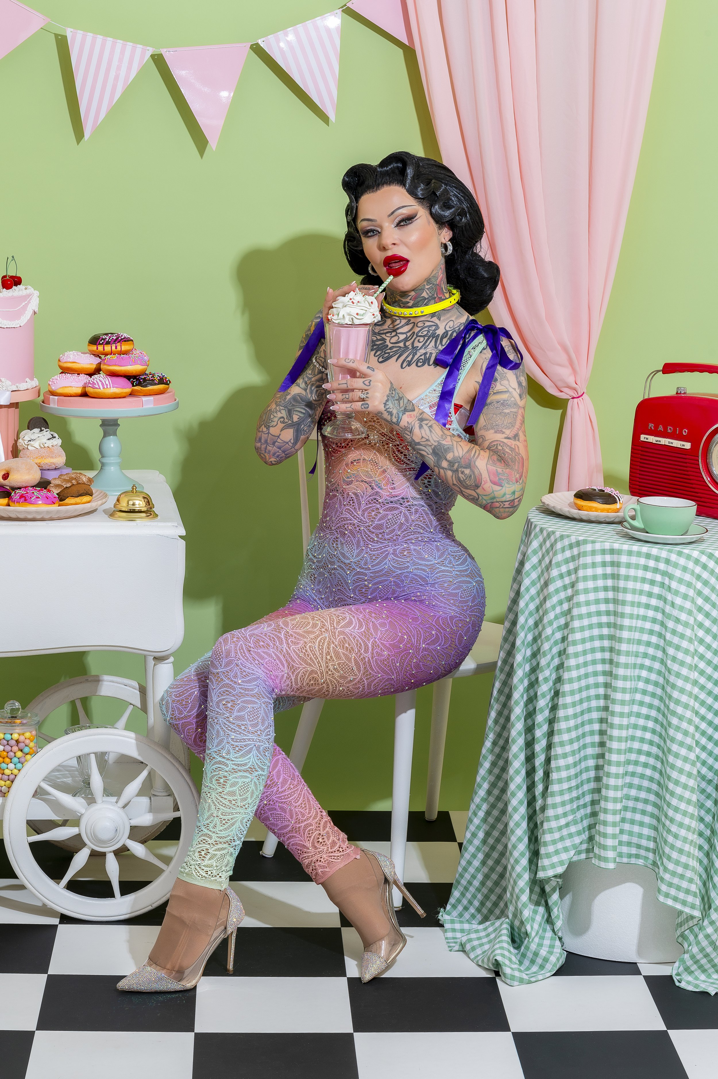 A tattooed woman in glittery lace clothing and high heels sits in a retro 1950s styled room with pastel colors, holding a milkshake with whipped cream and a striped straw. The room has a black and white checkered floor, a table with a checkered table