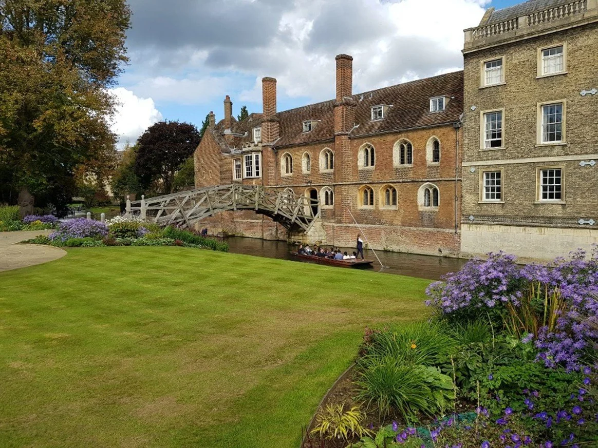 Historic brick buildings along a canal with a small arched bridge and a punt boat carrying passengers, surrounded by well-maintained lawn and colorful flower beds.