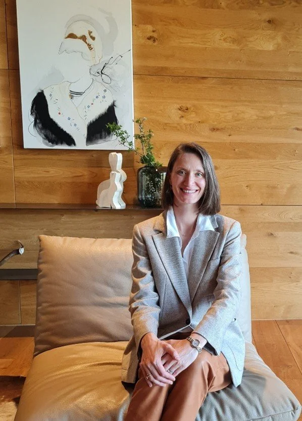 A woman sitting on a beige couch with a wooden wall background, smiling, wearing a light grey blazer and tan pants. Behind her is a painting of a woman with glasses, and decorative vases with greenery.
