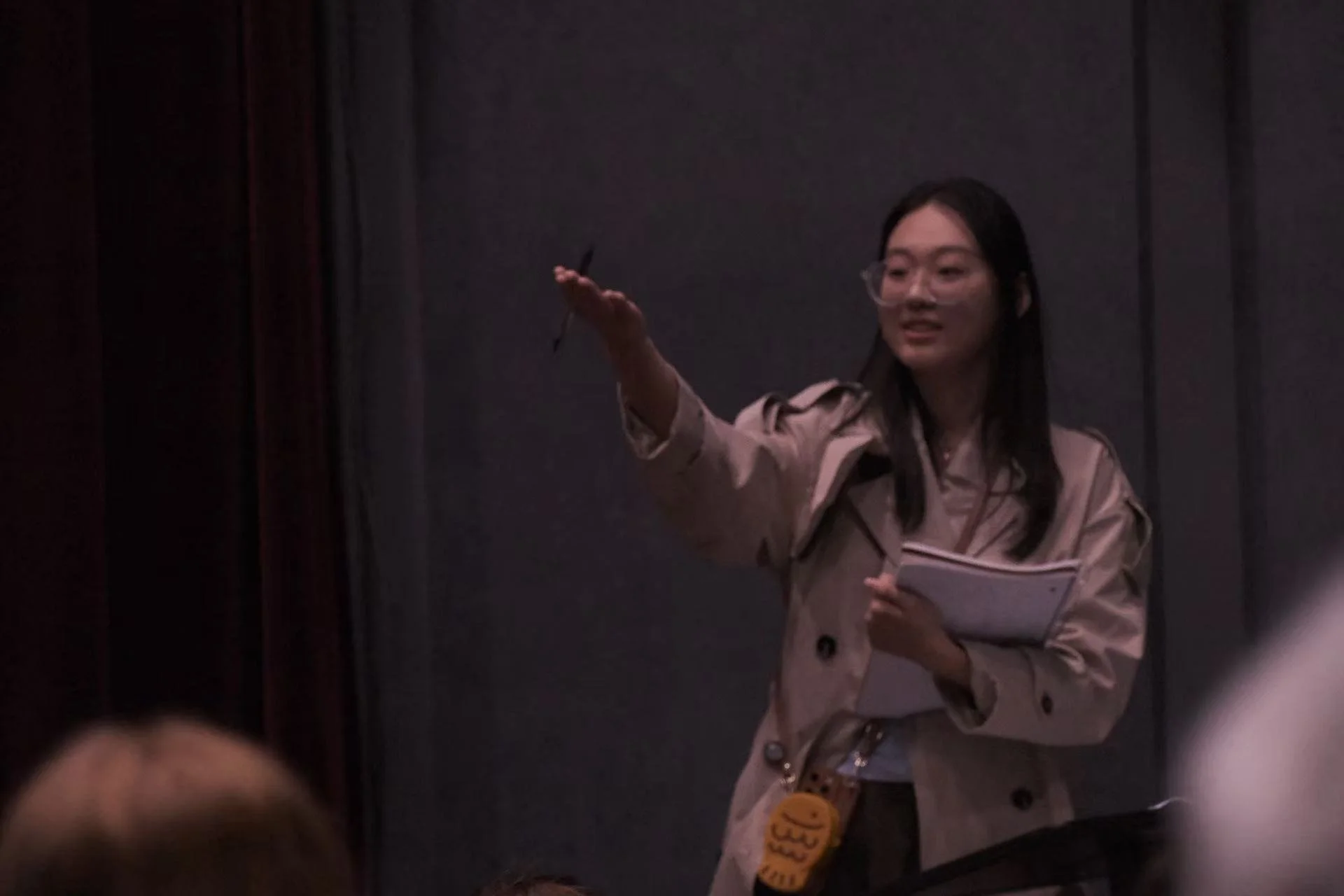 A woman standing in front of a dark background, holding a notepad, and gesturing with her hand while speaking or giving a presentation.