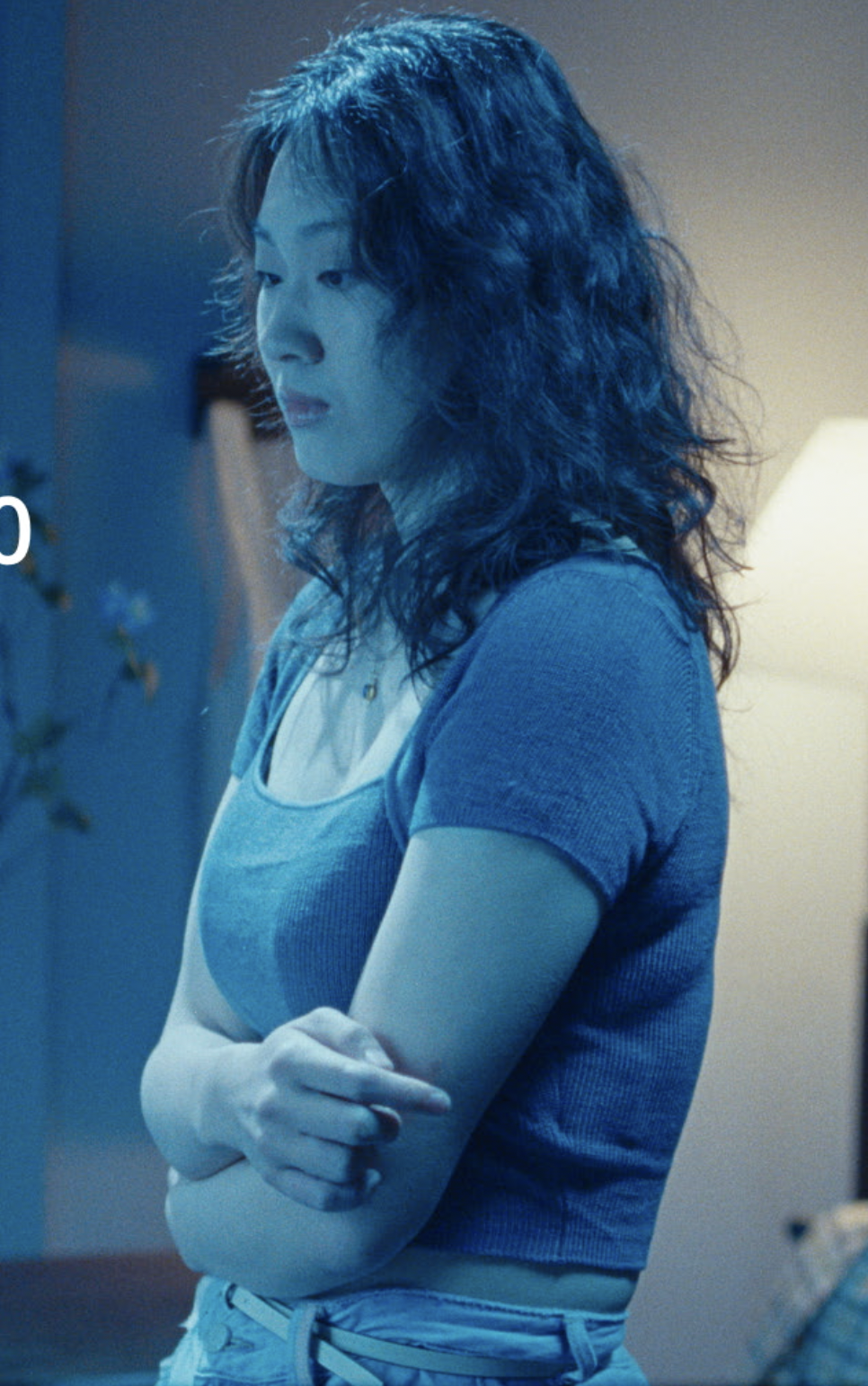 A young woman with wavy dark hair and light skin stands with arms crossed, looking down thoughtfully in a dimly lit room.