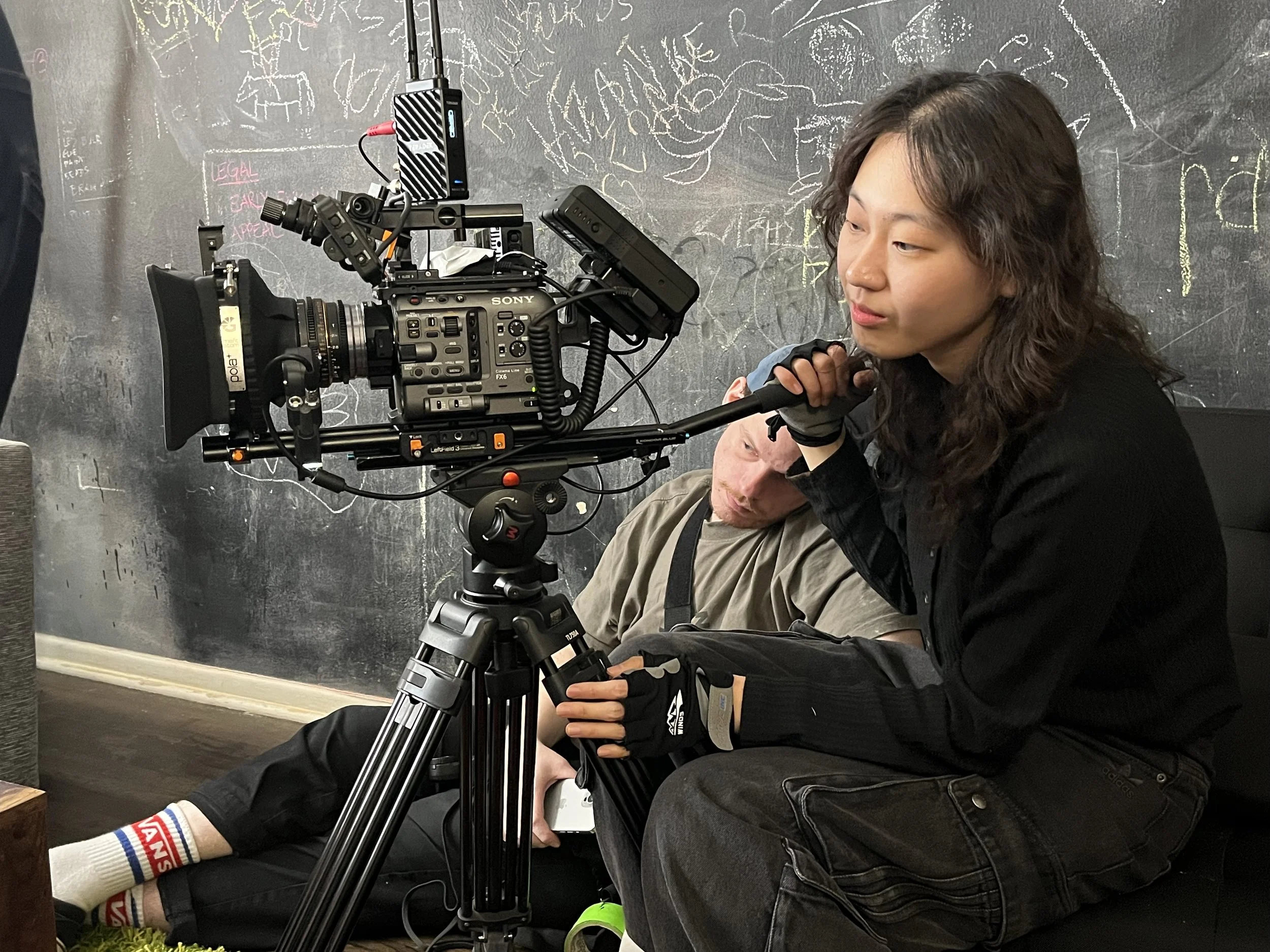 A woman operating a professional film camera on a tripod, sitting beside a man with a camera bag, in front of a blackboard with chalk drawings and writing.