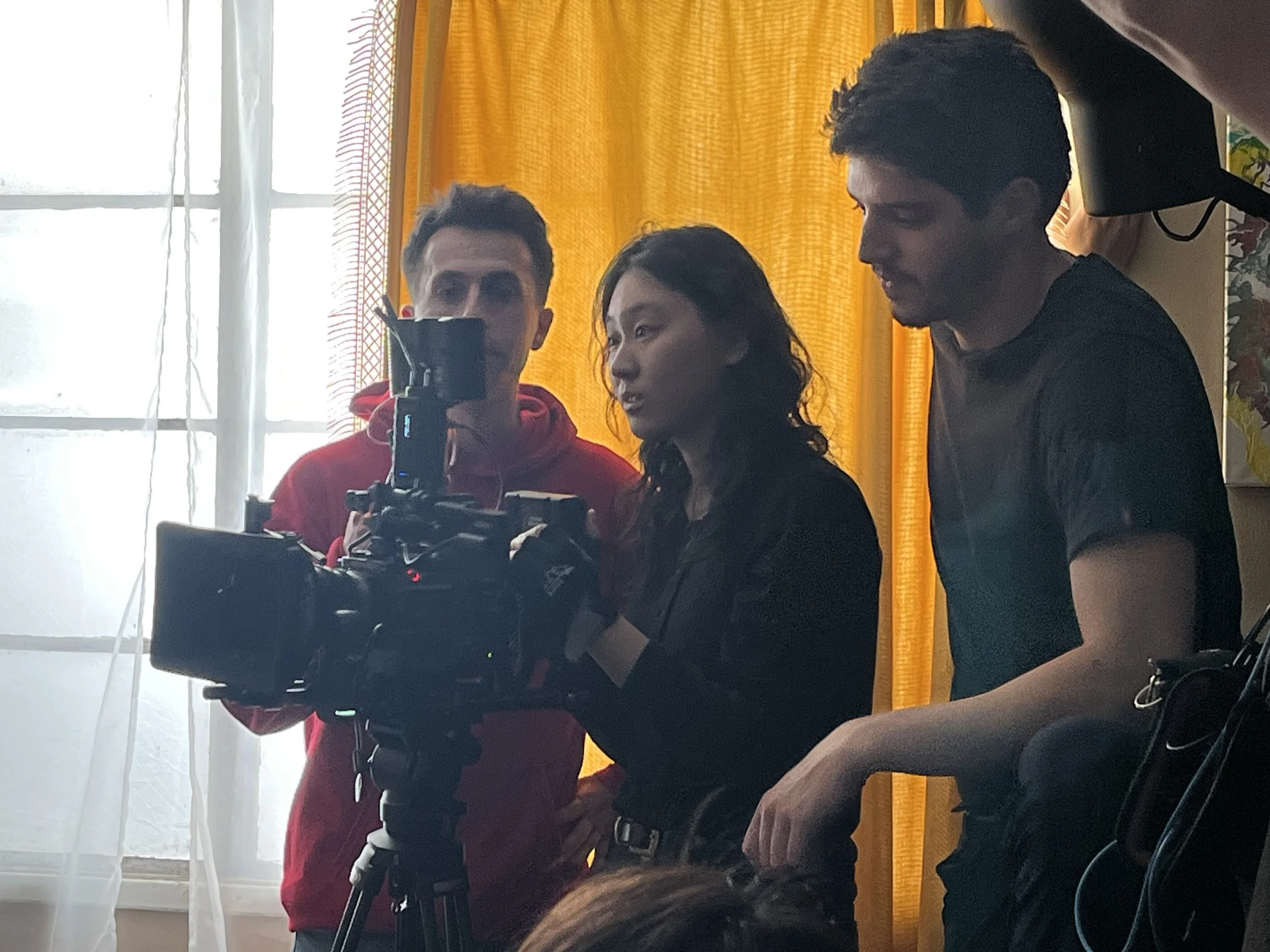 Three people working with a camera on a film set, indoors with a yellow curtain and bright window in the background.