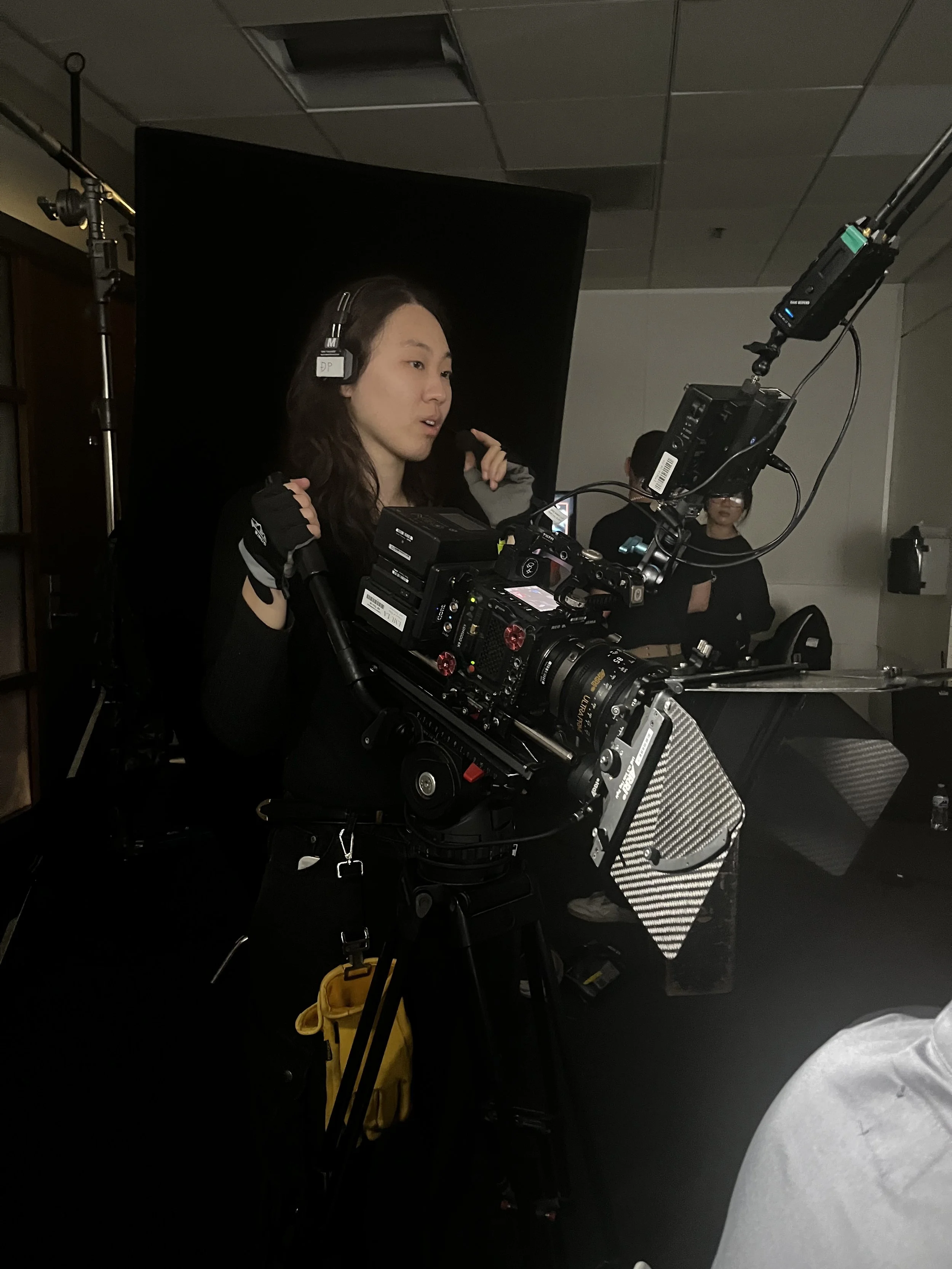 A woman operating a professional film camera during a shoot, with crew members and equipment visible in a dimly lit studio.