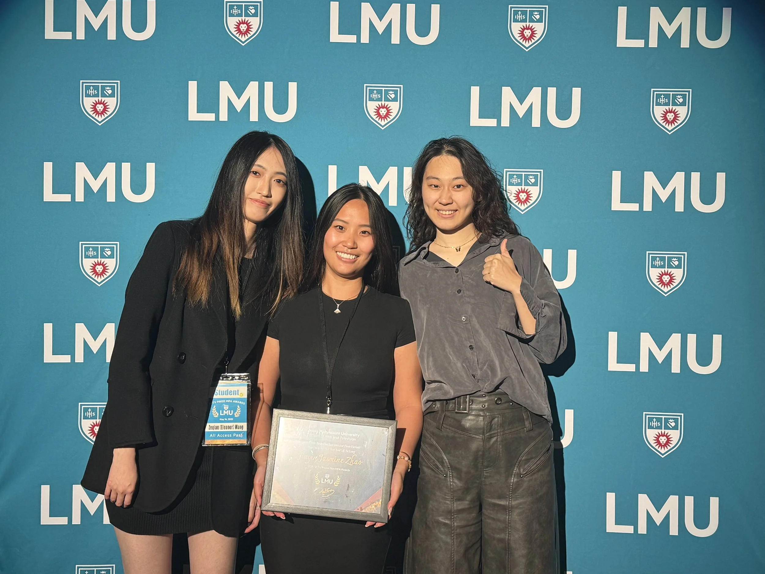 Three young women standing in front of a blue LMU backdrop, two of whom are smiling and one giving a thumbs up, one woman holding a certificate.