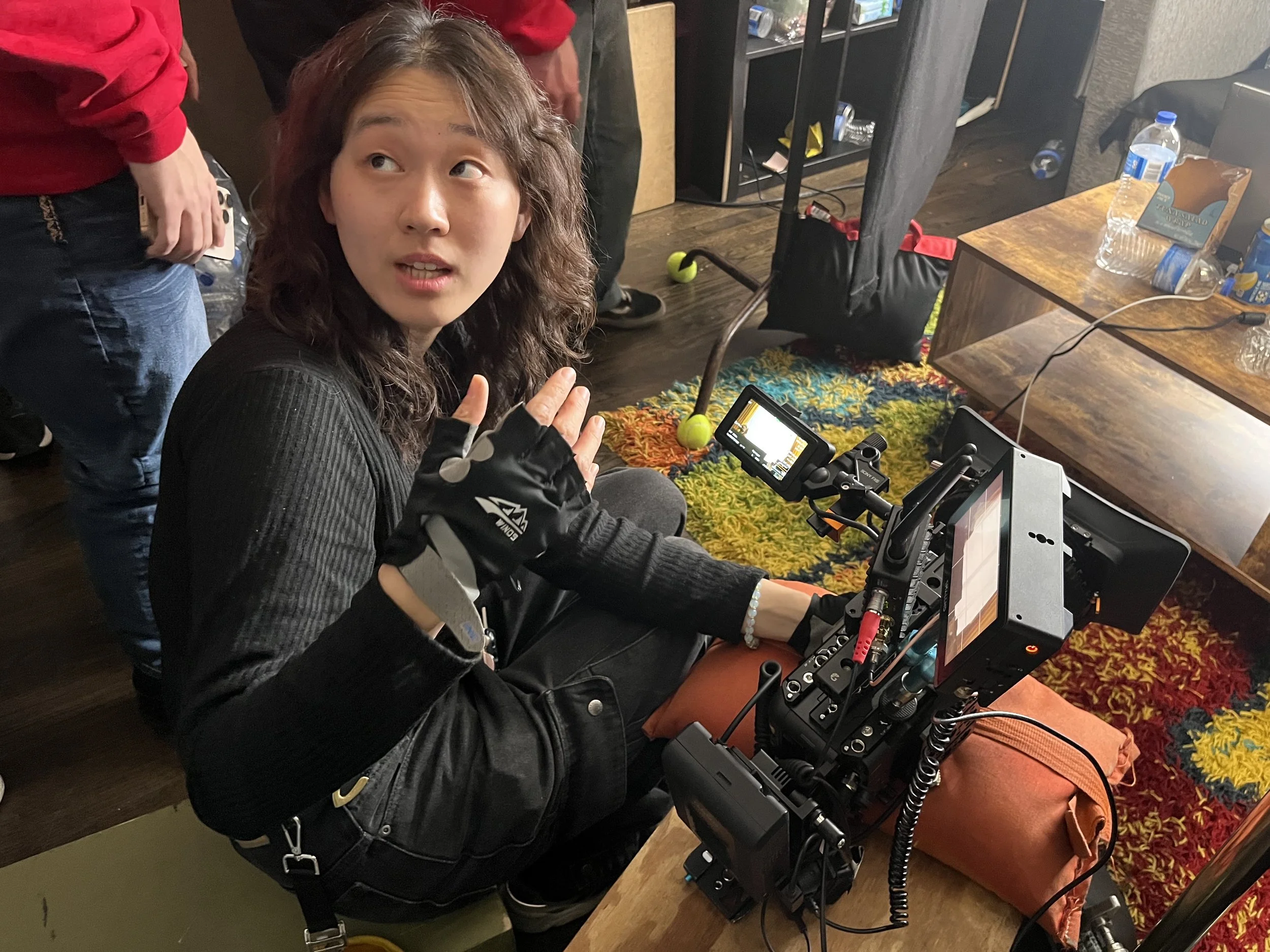 A woman sitting on a cushion operating a professional camera setup, with a woman in a red hoodie visible in the background, and a table with water bottles and snacks nearby.