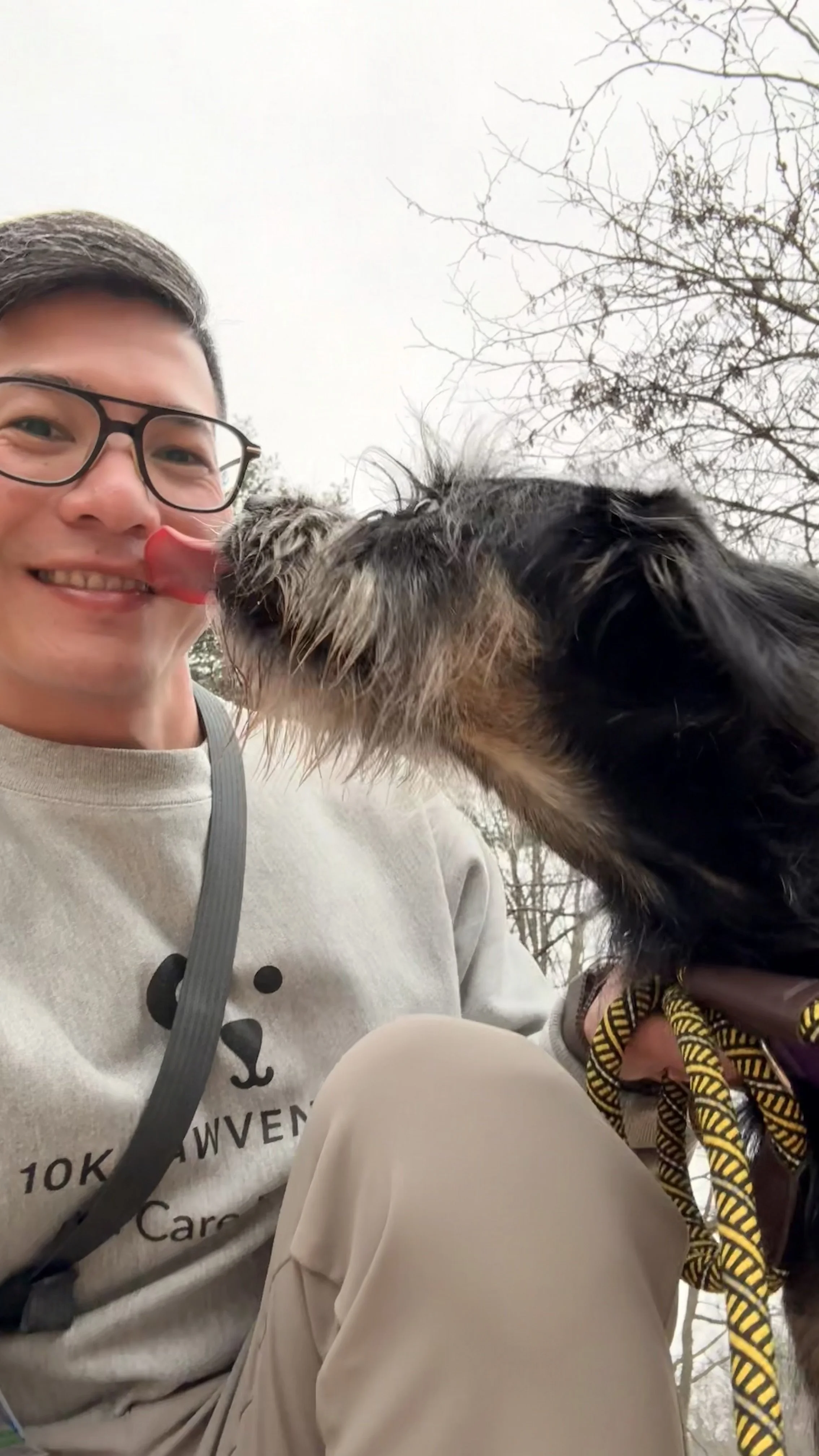 A young man with glasses smiling and showing his teeth, wearing a light gray sweatshirt with a bear face design, holding a scruffy black and gray dog that is licking his nose outdoors in a park with bare trees.