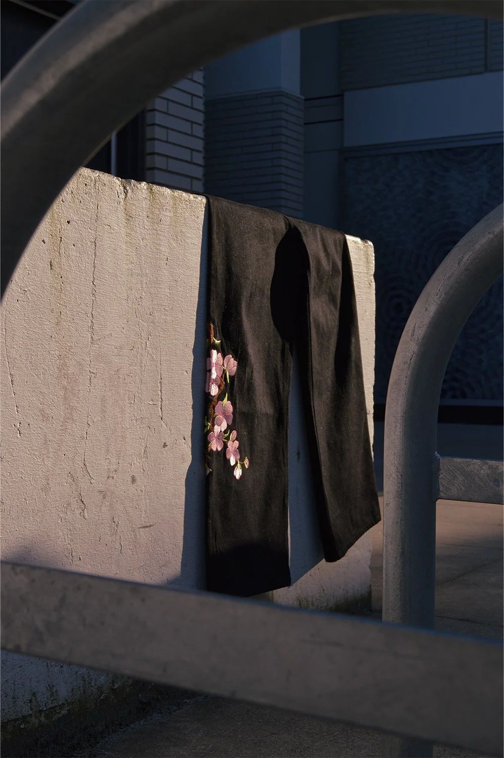 A pair of black pants with pink embroidered flowers hanging on a white concrete wall, framed by metal railings, with a modern building in the background.
