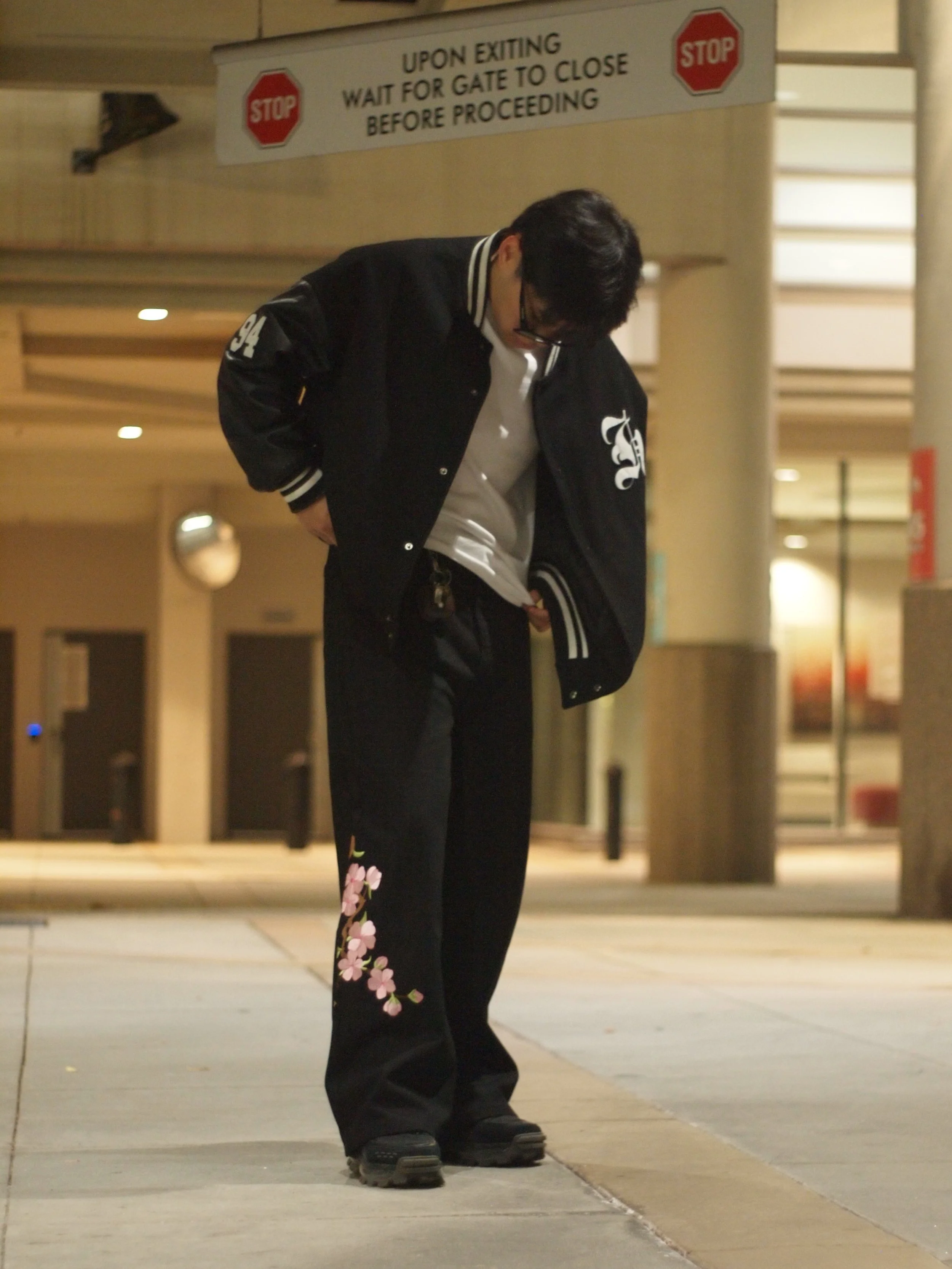 A person standing outside at night, looking down, wearing black pants with pink flower embroidery, a black jacket, and glasses, beneath a sign that reads, 'Upon exiting, wait for gate to close before proceeding.'