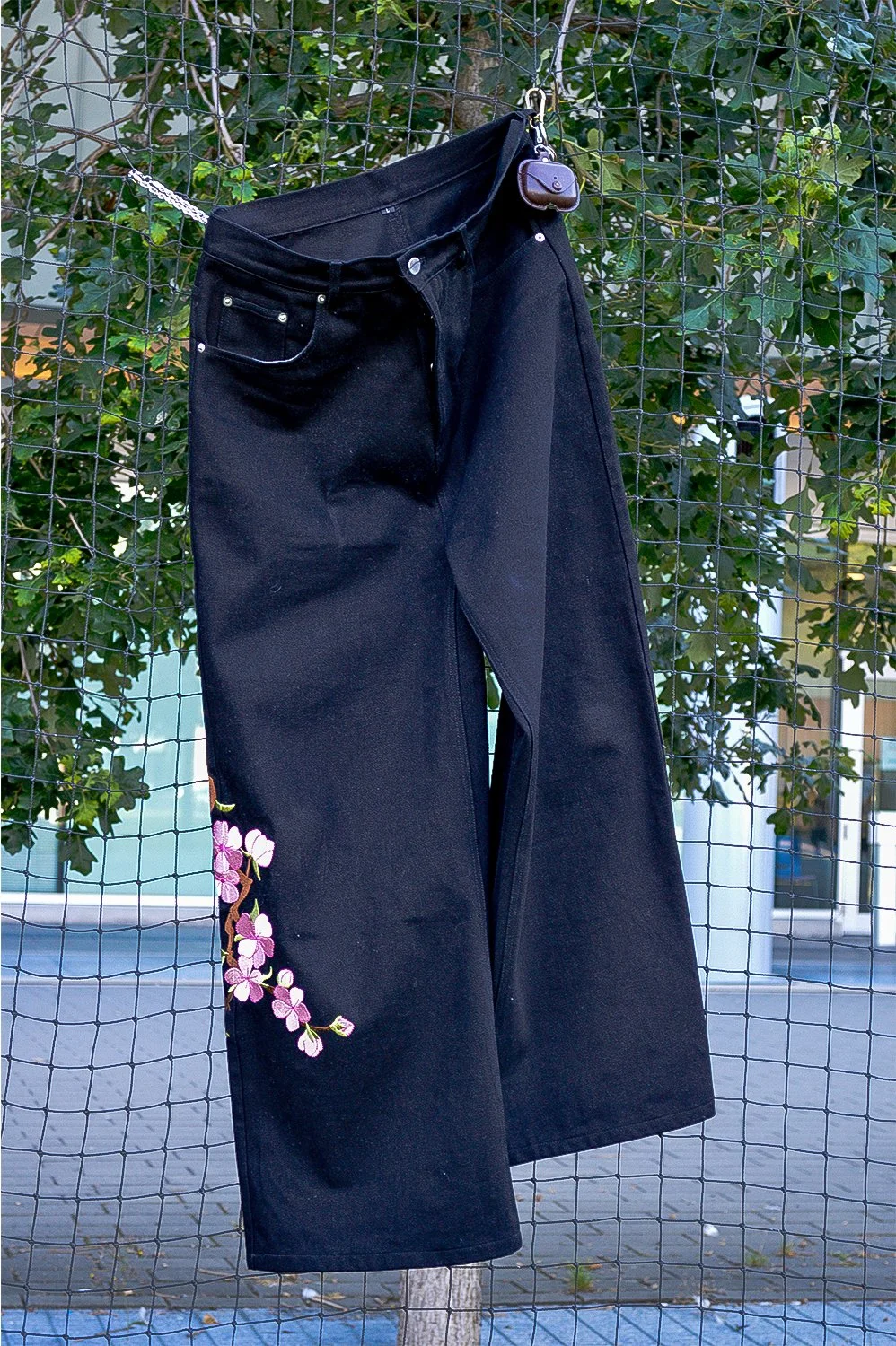 Black jeans with floral embroidery hanging on a wire fence, with green foliage in the background.