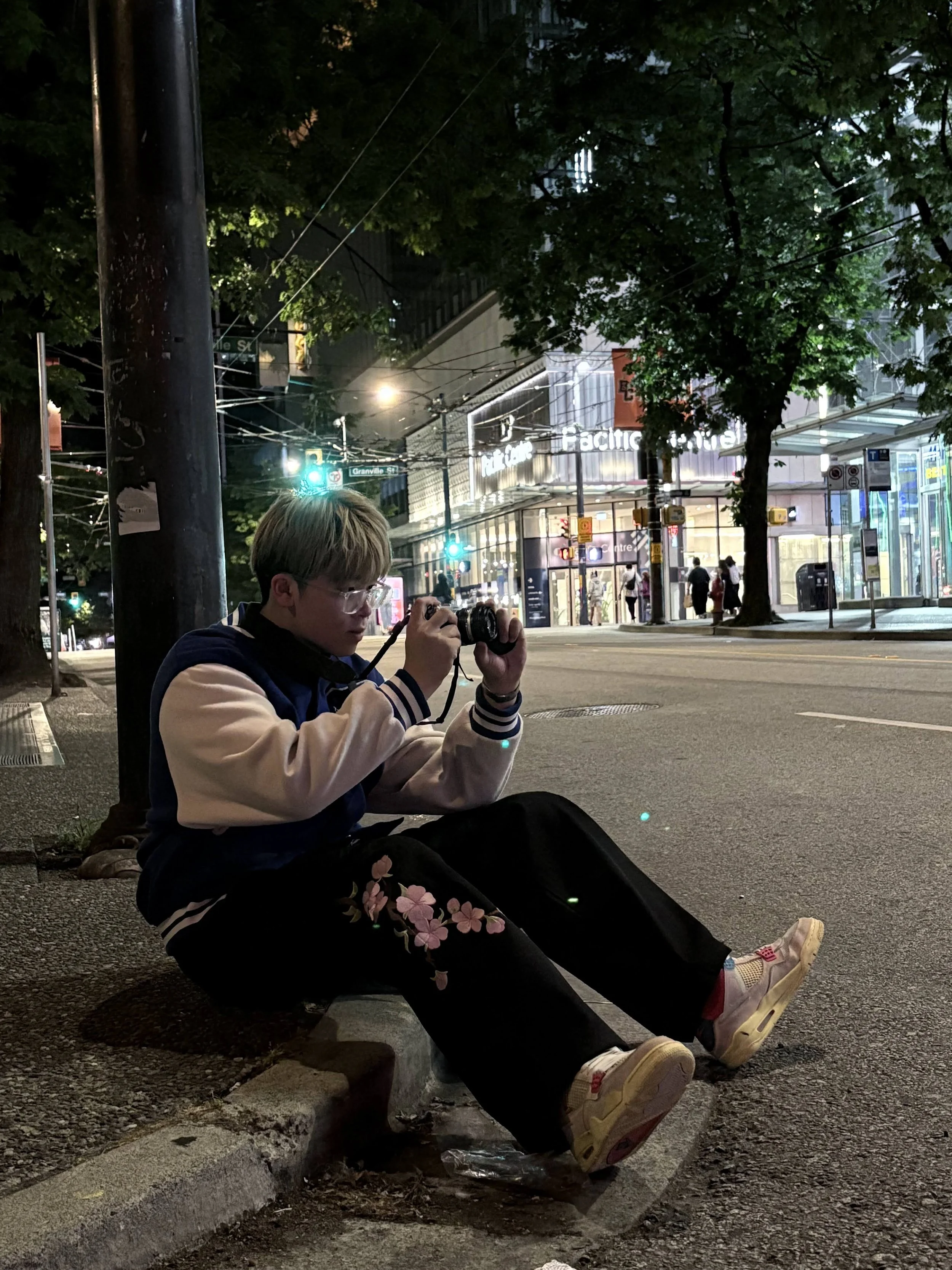 A person sitting on a curb at night, taking a photo with a camera, with city lights and storefronts in the background.
