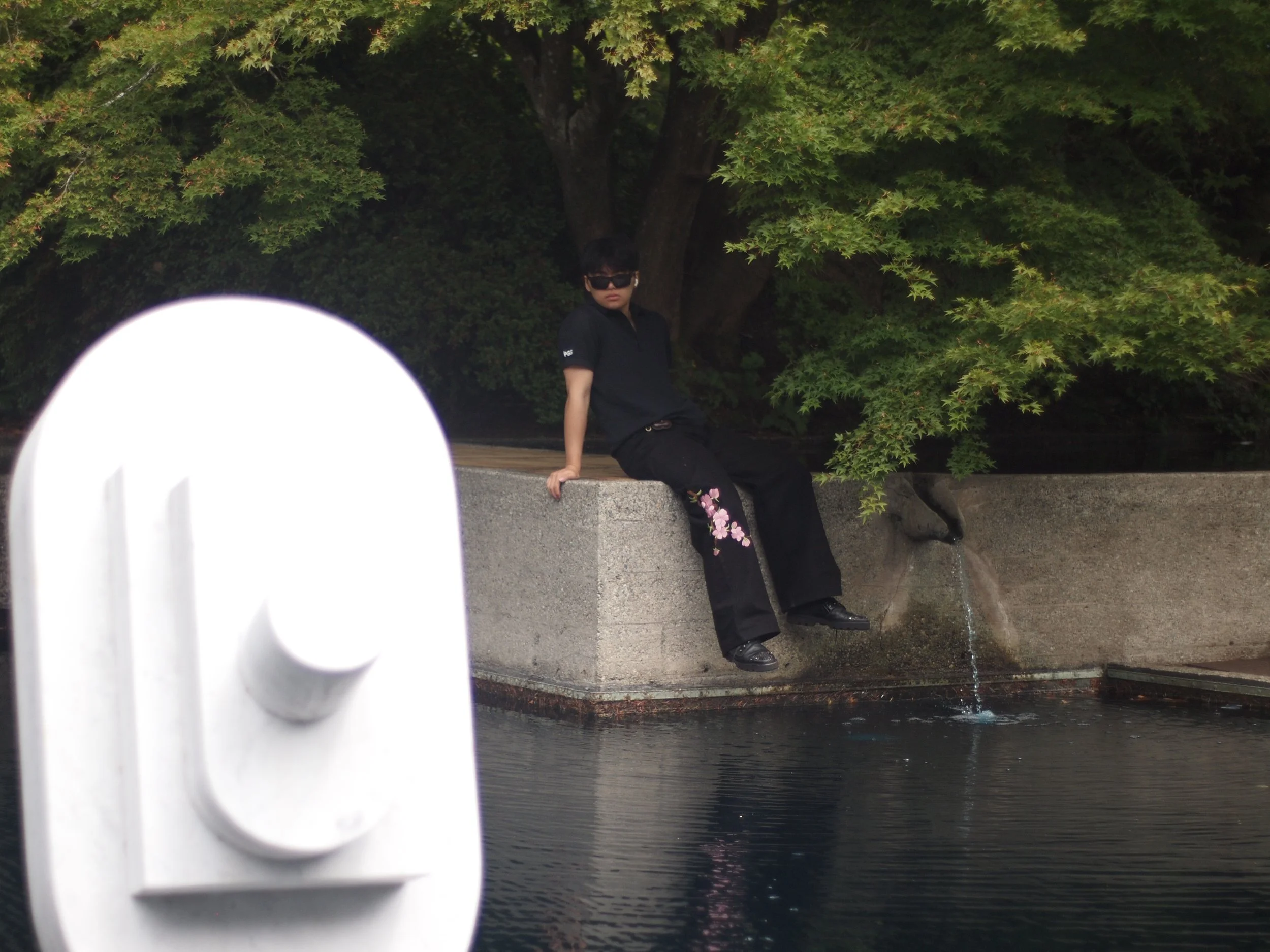 A woman in black clothing and sunglasses sitting on a concrete ledge by a pond, with trees and greenery in the background, and a fountain nozzle in the foreground.