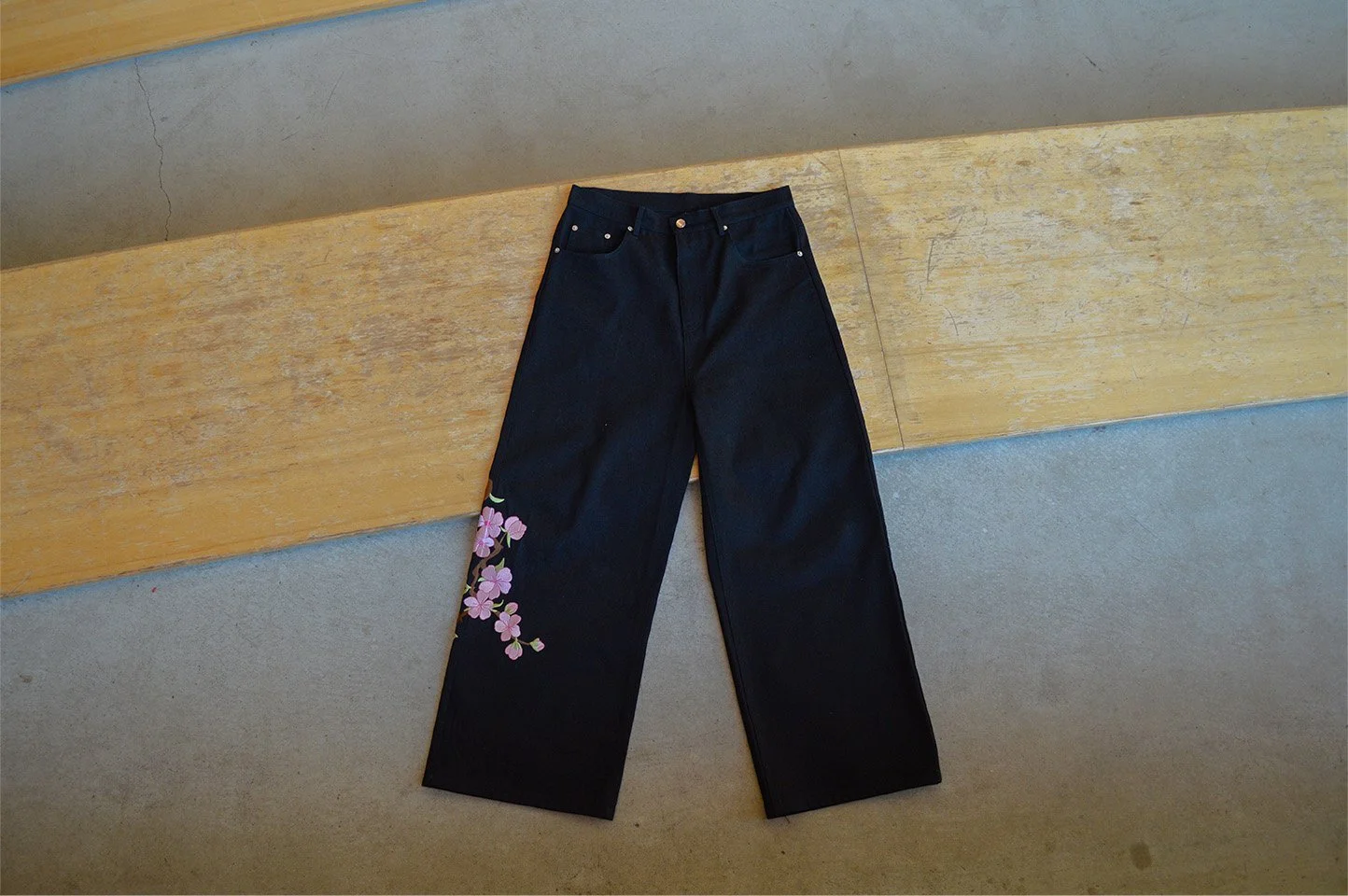 Black wide-leg jeans with pink floral embroidery on the lower left leg, laid flat on a wooden surface.