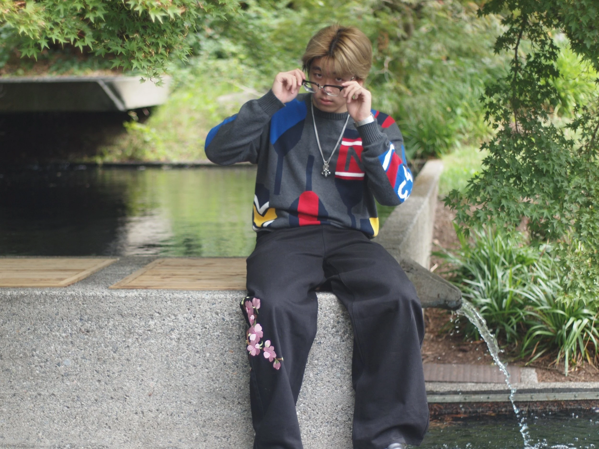 A young person with blond hair and glasses sits on a concrete ledge near a water feature, adjusting their glasses and looking at the camera, surrounded by greenery.