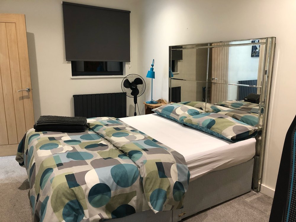 A bedroom with a bed covered in a multicolored geometric patterned duvet and matching pillows. The bed has a mirrored headboard, and there is a small wooden side table with a blue desk lamp, a fan, and some items on it. A window with a closed dark sh