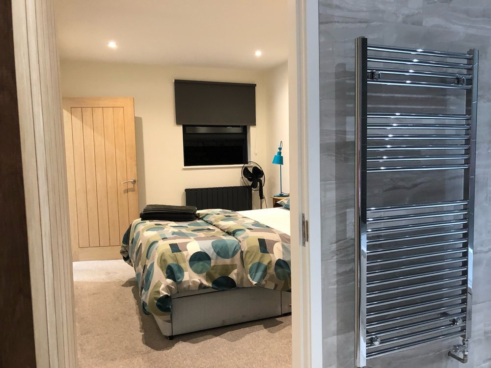 A bedroom with a bed covered in a colorful geometric duvet, a small bedside table with a blue lamp, a window with a dark shade, and a towel radiator in a bathroom.