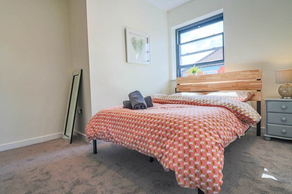 Bedroom with a bed covered in a pink and beige geometric patterned comforter, a gray nightstand with a lamp, a window with a plant on the sill, and a picture on the wall, with a full-length mirror leaning against the wall.