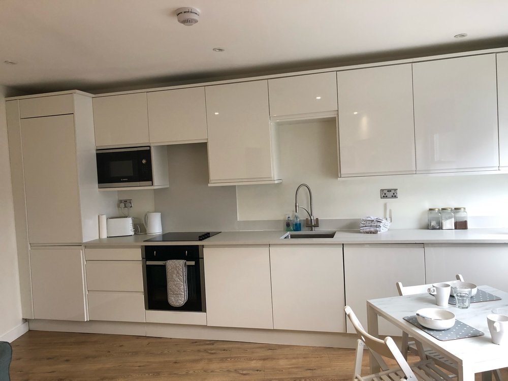 Modern white kitchen with built-in microwave, stovetop, oven, stainless steel sink, and minimal decor. Dining table with white chairs and tableware in the foreground.