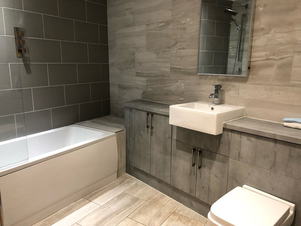 Modern bathroom with gray tiled walls and floors, a white bathtub with a glass shower partition, a small wall-mounted sink, a large mirror, gray cabinetry, and a toilet.