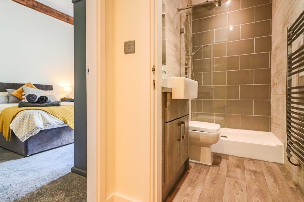 View of a bedroom with a bed featuring yellow and white bedding, towels on the bed, and part of an adjacent bathroom with a walk-in shower, toilet, sink, wooden vanity, and towel radiator.