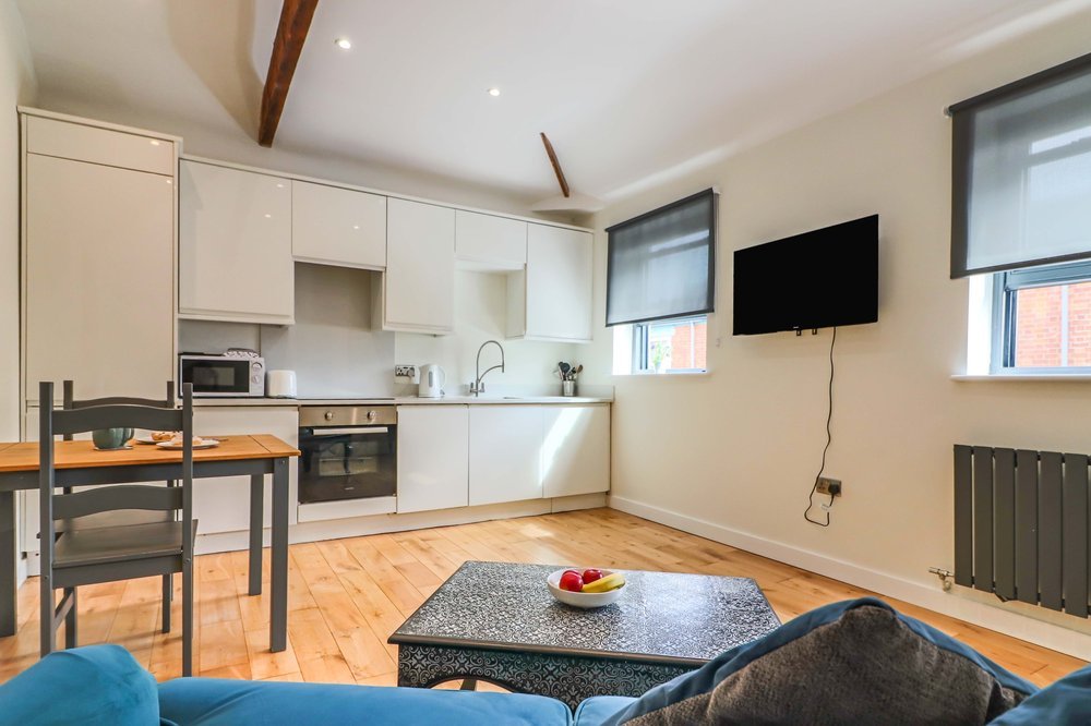 Living room with open kitchen, white cabinets, a wall-mounted TV, hardwood floor, windows with blinds, and a blue sofa with a coffee table holding a bowl of apples.