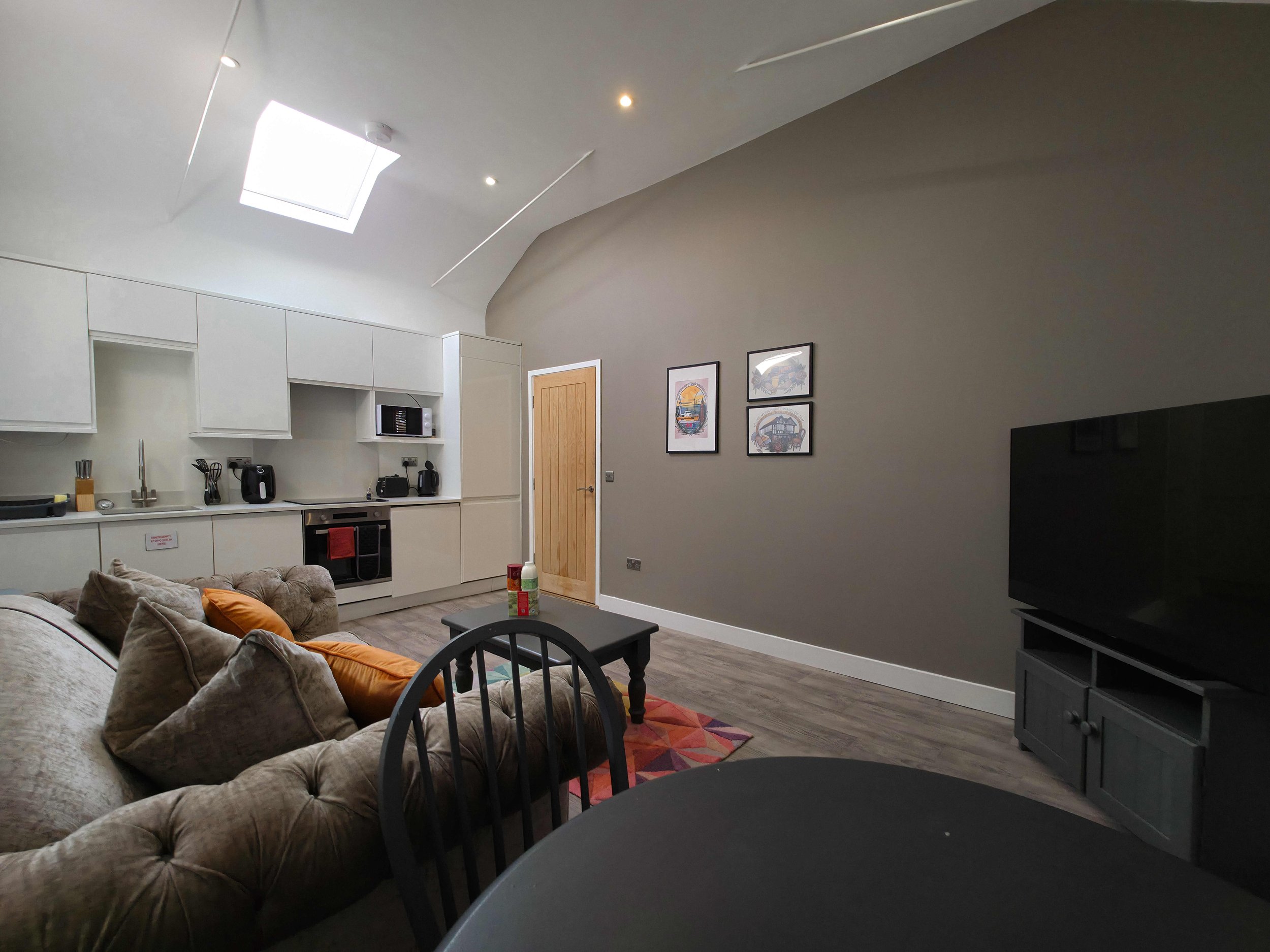 Living room with beige sofa, black chairs, small black coffee table, wall art, TV on gray stand, kitchen area with white cabinets, microwave, and small appliances, ceiling skylight, and gray walls.
