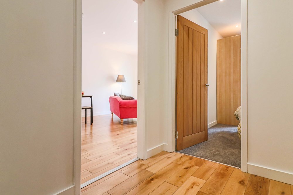Interiors of a home showing a hallway with doorways leading to a living room with a red sofa and a side table, and a bedroom with a bed and nightstand. The floors are wood and carpet.