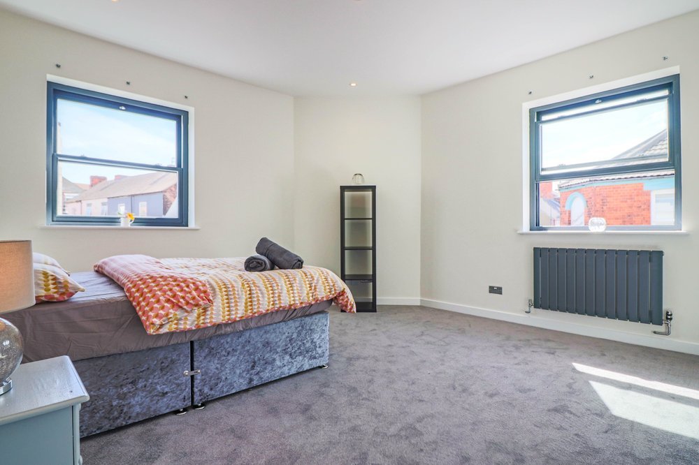 Bedroom with two windows, a bed with colorful blanket and pillows, a small bedside table, a black ladder shelf, and a radiator.