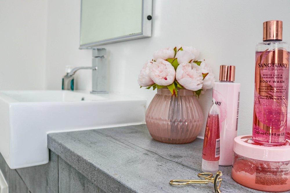 Bathroom countertop with pink flowers in a vase, skincare products, a mirror, and a sink.