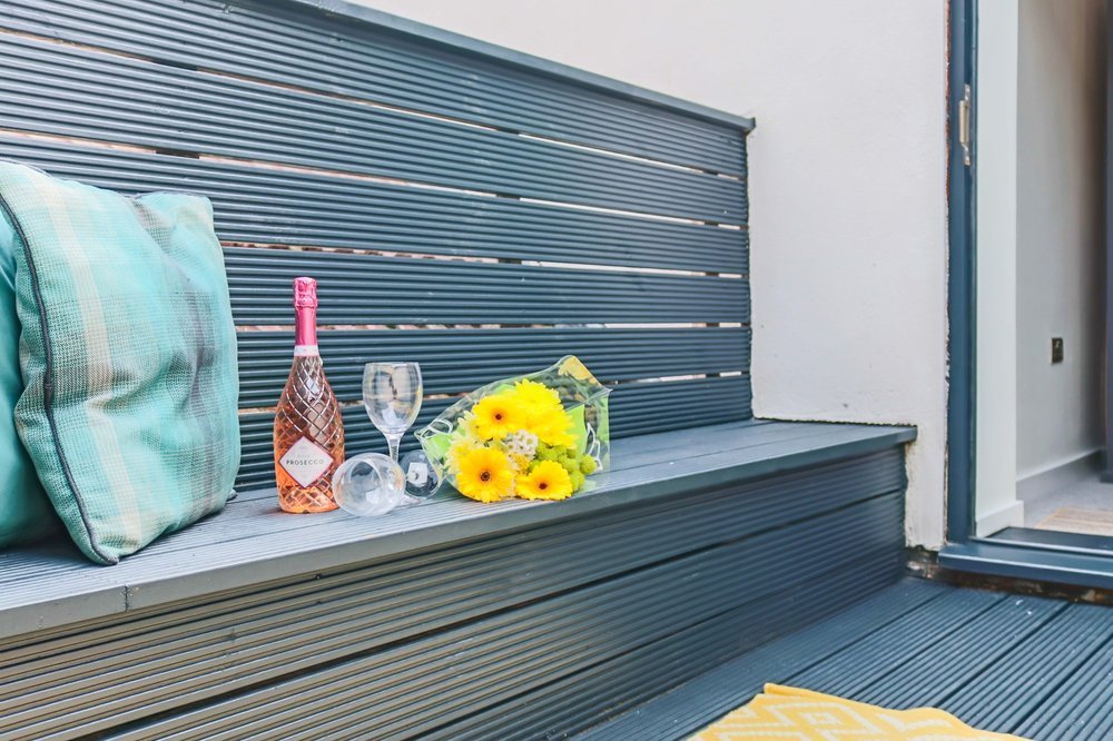 A bouquet of yellow flowers, a bottle of rosé wine, two empty wine glasses, and a pillow placed on a gray outdoor bench near a door.