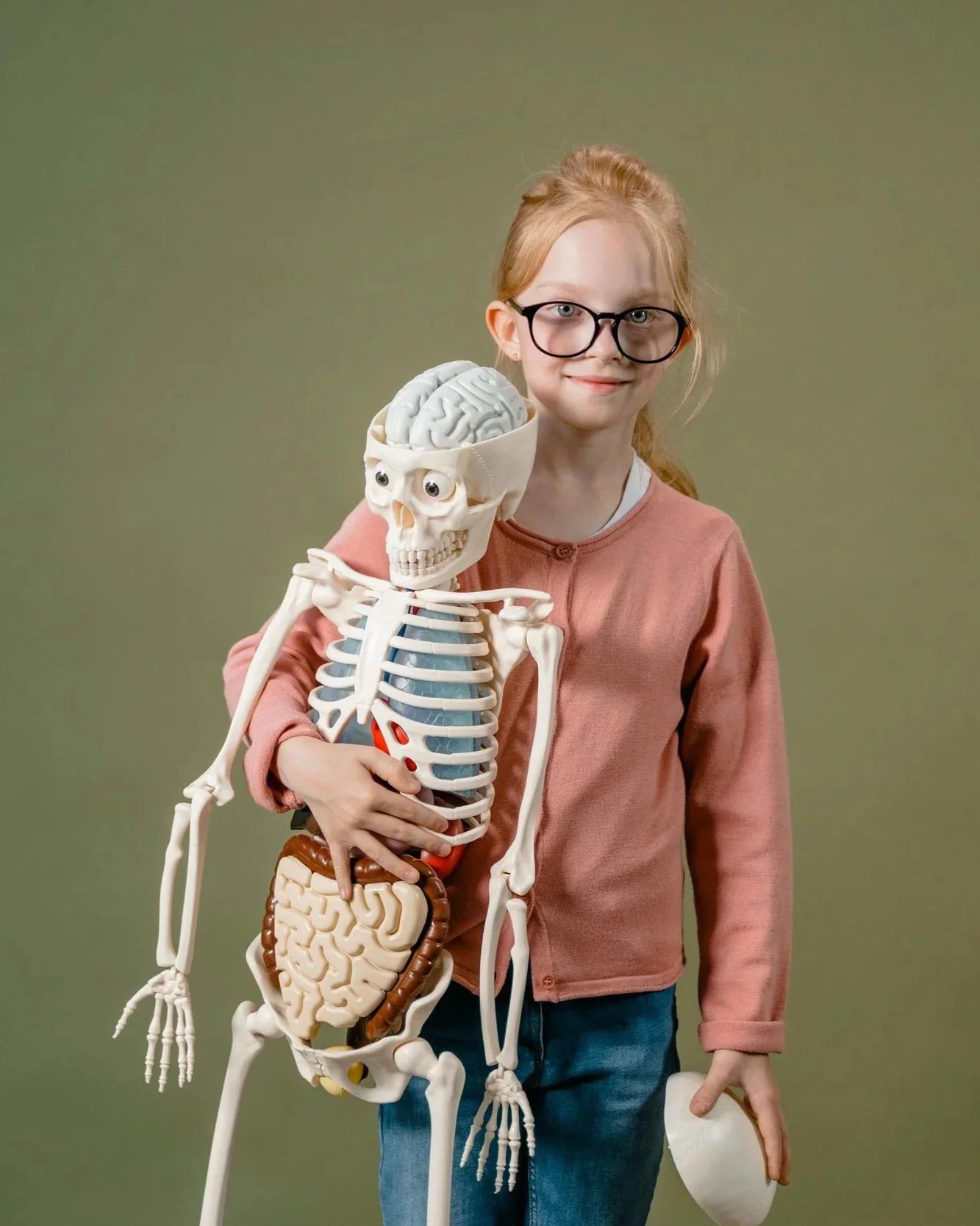 A young girl with red hair and glasses standing next to a plastic skeleton model, holding it around the waist and smiling at the camera. The girl is wearing a pink jacket and blue jeans, with a plain green background.