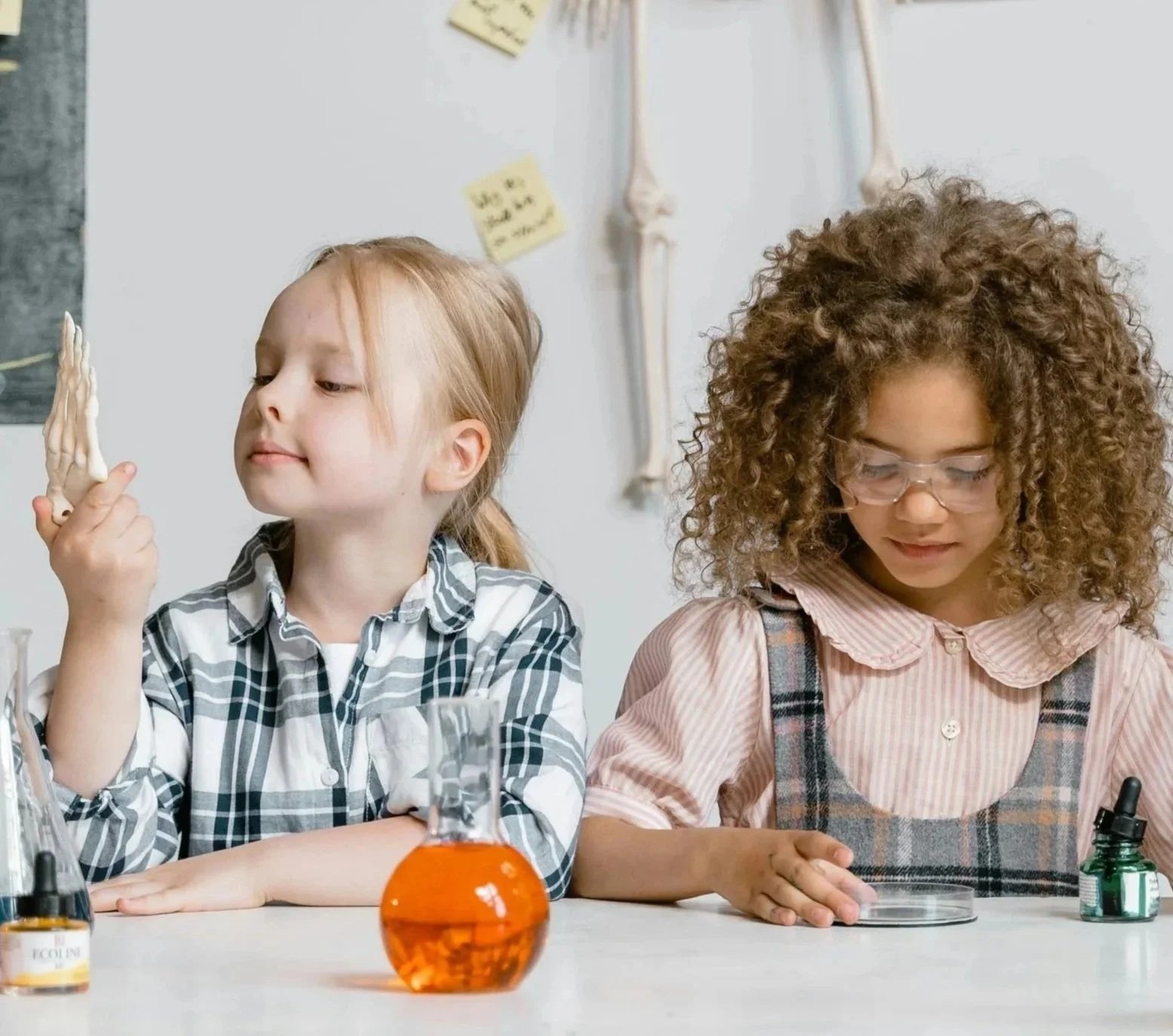 Two young girls sitting at a table in a science lab, with science equipment and posters on the wall behind them, engaging in a science activity.
