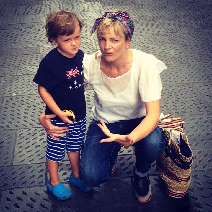 A woman with a colorful headscarf kneeling on one knee on a textured pavement, holding a young boy with curly hair wearing a navy blue shirt, striped shorts, and blue shoes. The woman is wearing a white t-shirt, jeans, and sneakers. She has a striped bag on the ground next to her. They are outdoors on a paved area.